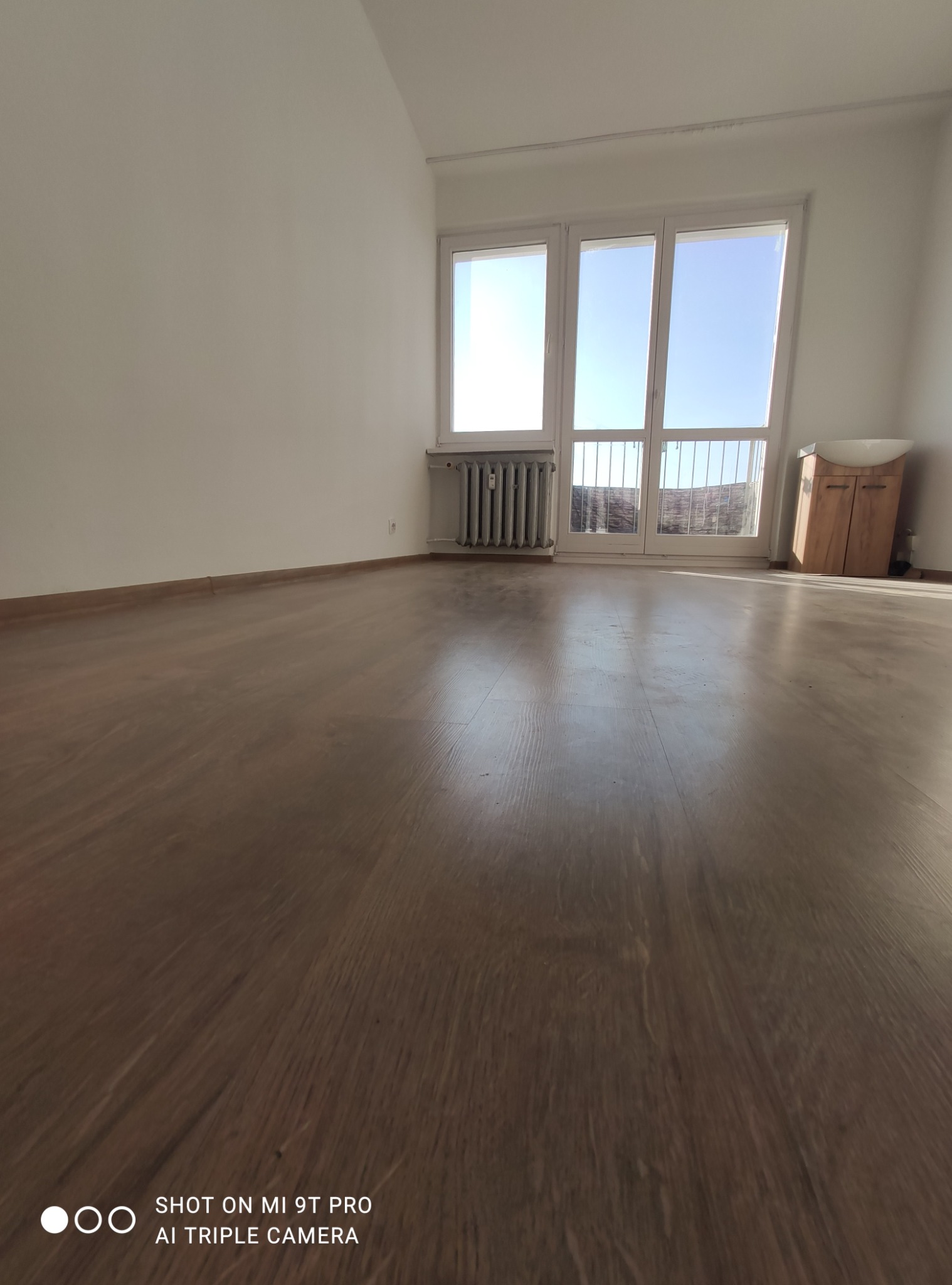 Puste, odświeżone wnętrze z drewnianą podłogą, białymi ścianami i grzejnikiem przy oknie balkonowym, gotowe do aranżacji.