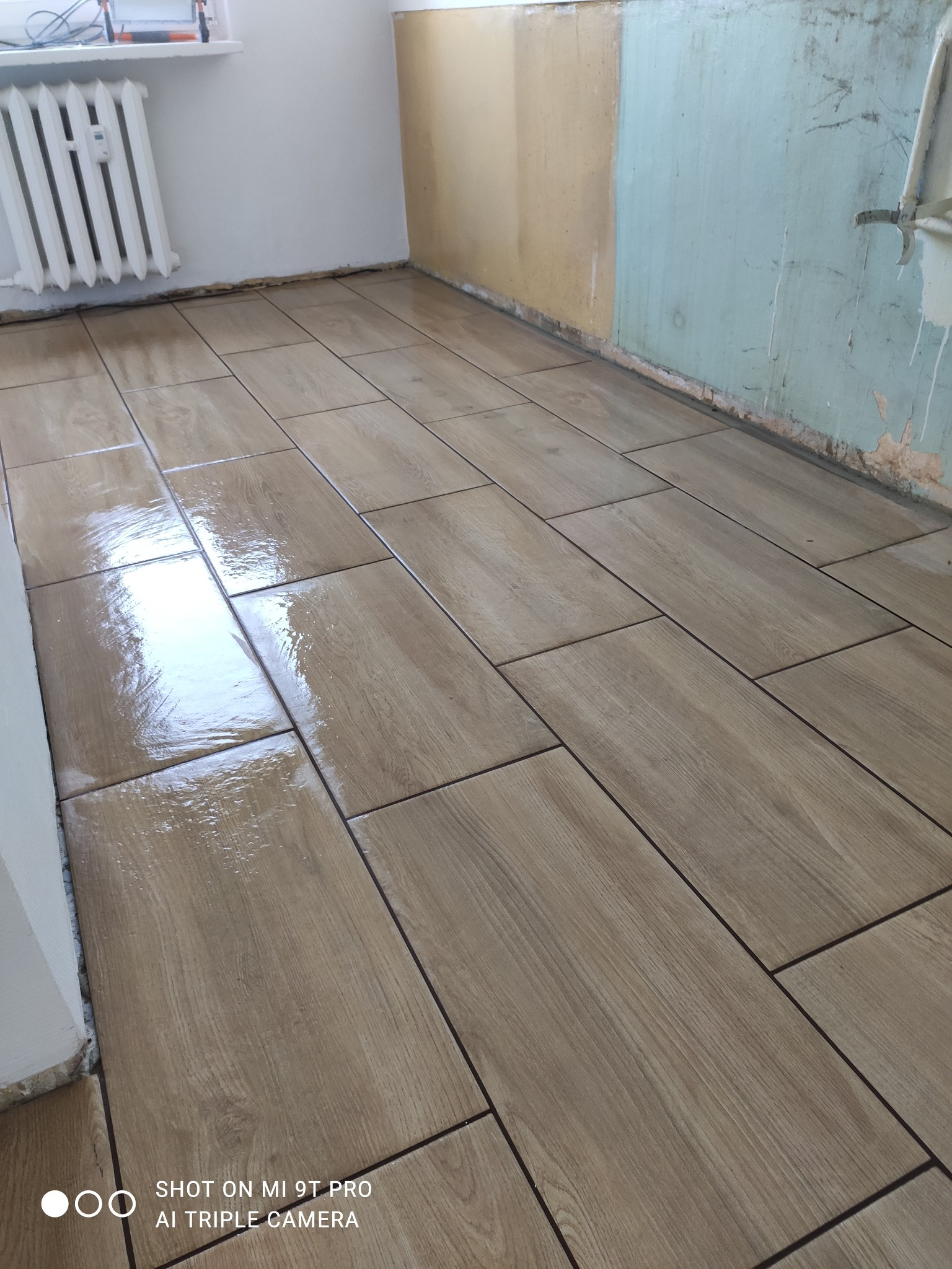 Ułożone, prostokątne płytki podłogowe imitujące drewno, z ciemną fugą. W tle fragment ściany w kolorze beżowym i błękitnym, oraz biały grzejnik.