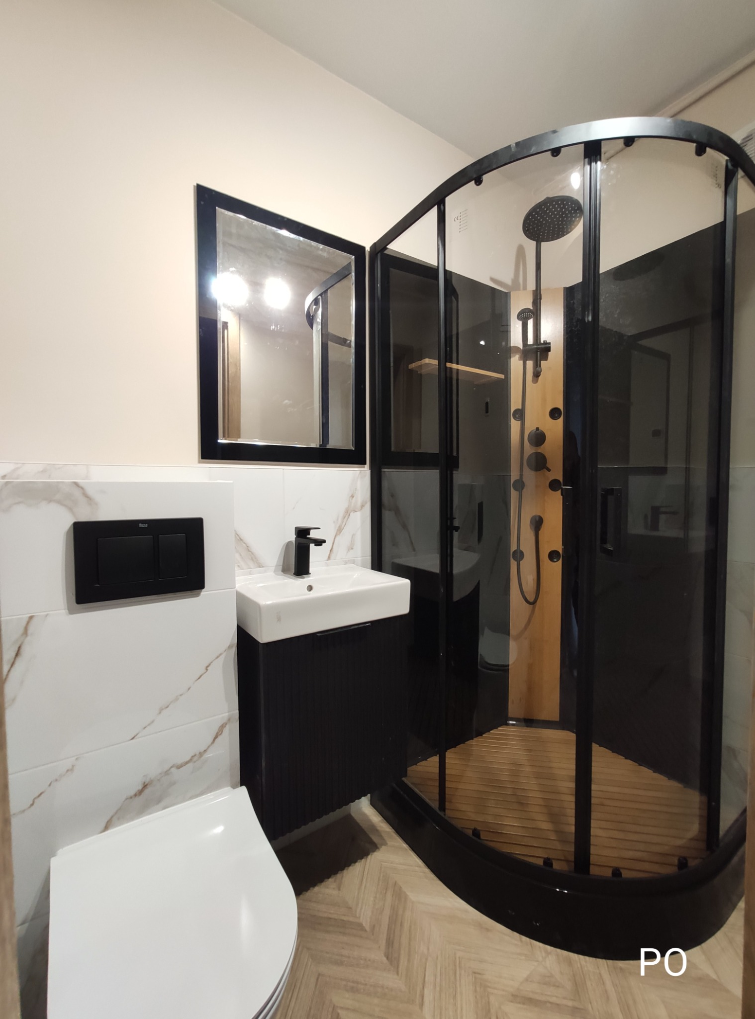 Nowoczesna łazienka po remoncie: kabina prysznicowa z drewnianym panelem, umywalka z czarną szafką i lustro w czarnej ramie. Białe płytki imitujące marmur.