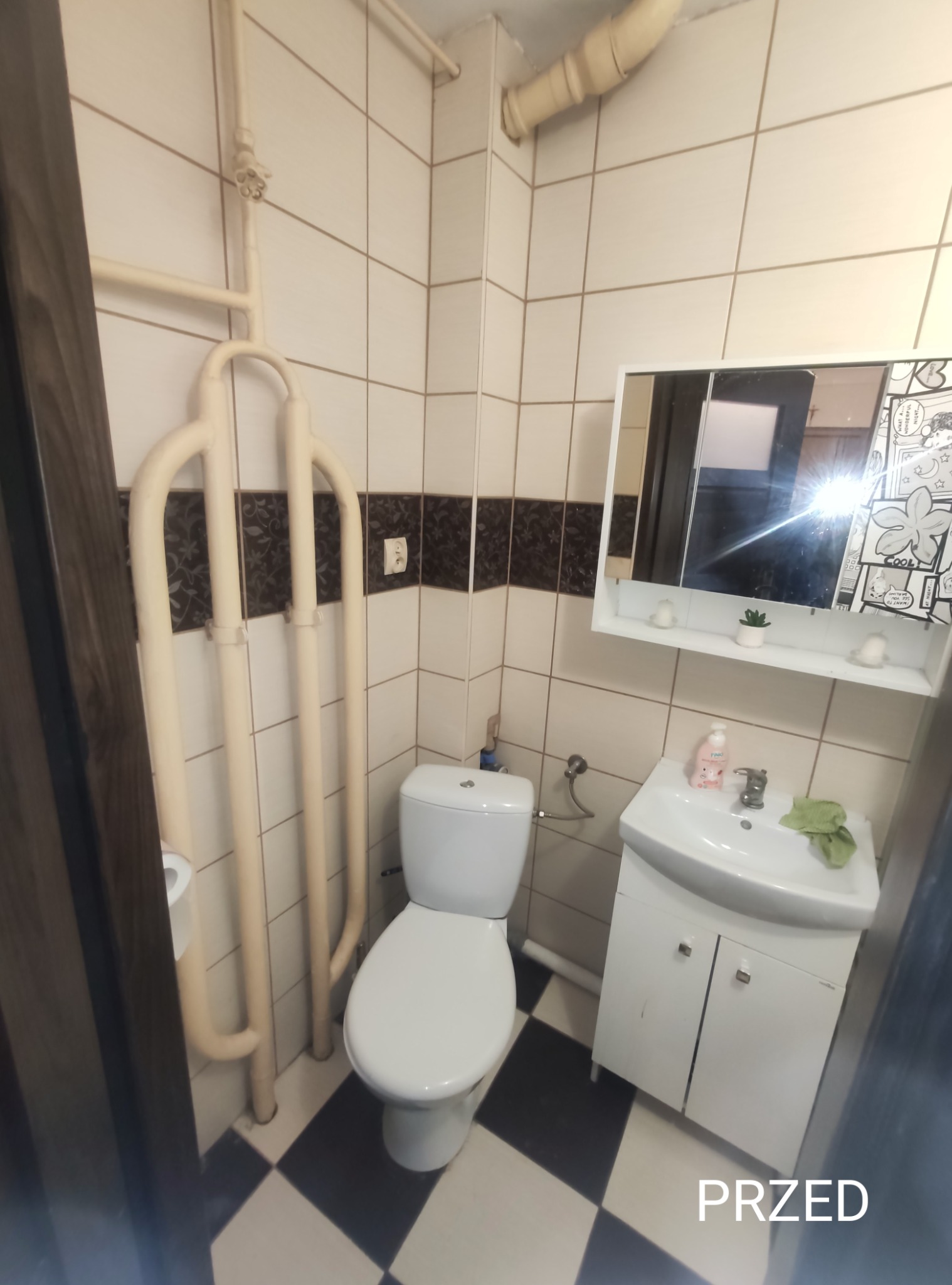 Zdjęcie łazienki z białą armaturą i czarno-białą podłogą w kratkę, z widocznym napisem 'PRZED', sugerujące stan przed remontem. Grzejnik drabinkowy w kremowym kolorze.