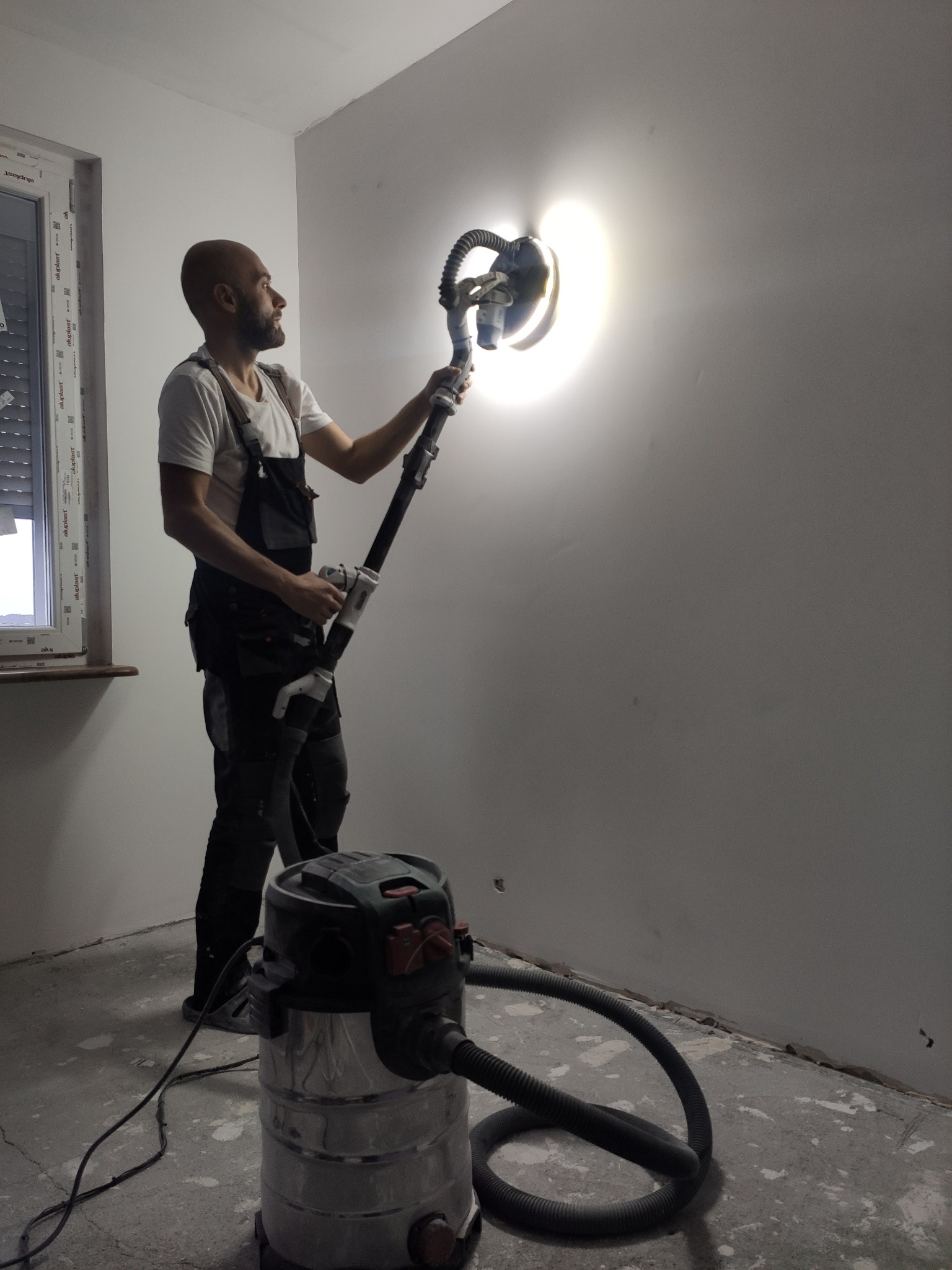 Szpachlowanie ściany szlifierką z odkurzaczem przez mężczyznę w kombinezonie roboczym. Widoczne okno z roletami i fragment podłogi w trakcie remontu.