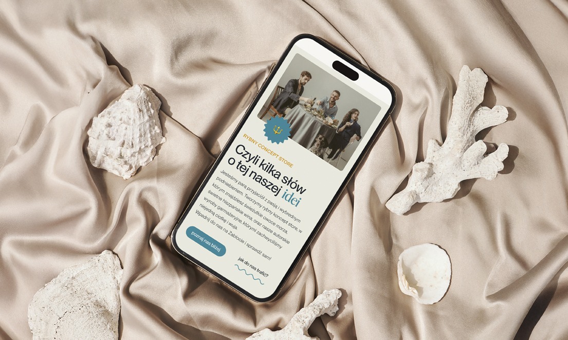 Smartfon wyświetla mobilną wersję strony internetowej z ofertą rybnego concept store, otoczony dekoracyjnymi elementami morskimi na satynowym tle.