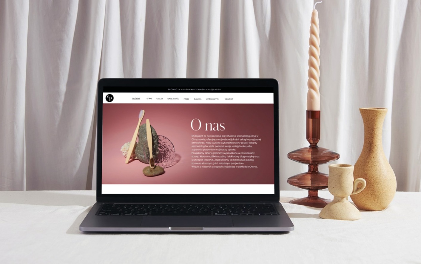 Laptop wyświetlający stronę internetową gabinetu stomatologicznego z zakładką 'O nas', obok świeca w szklanym świeczniku i beżowe wazony na białym obrusie.