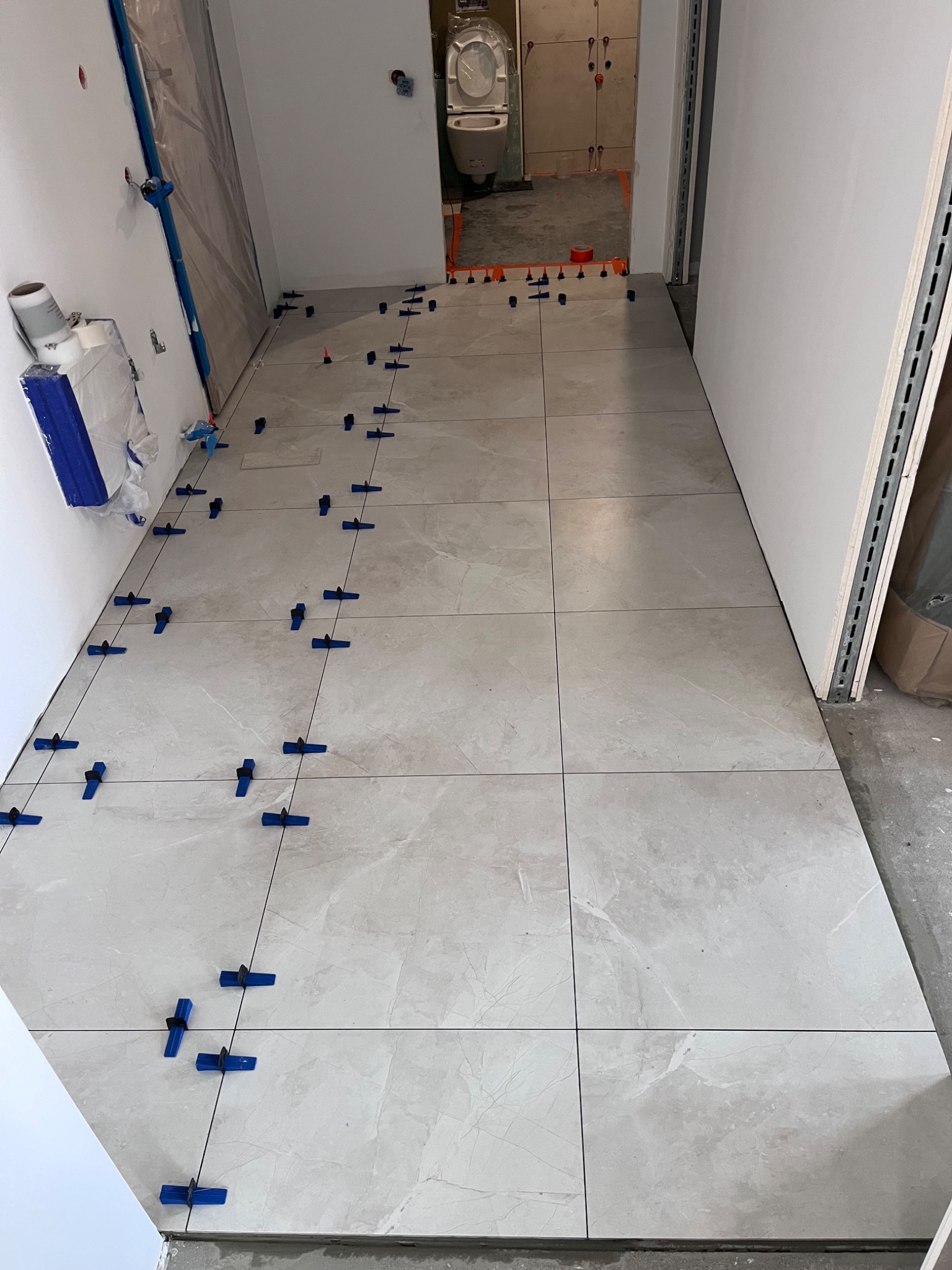 Układanie jasnych, marmurkowych płytek podłogowych w korytarzu z użyciem niebieskich klipsów poziomujących i krzyżyków dystansowych; widoczny fragment toalety w tle.