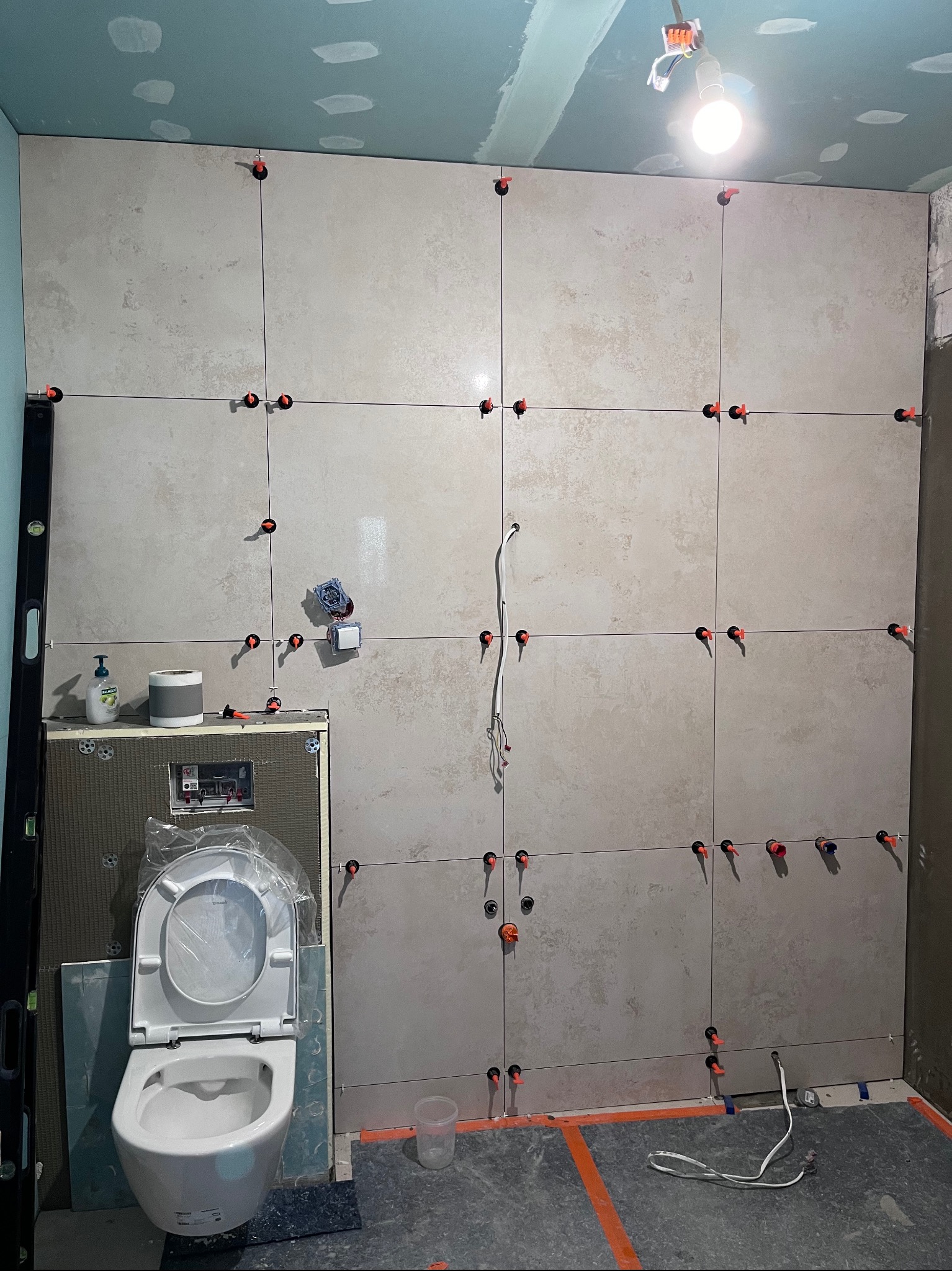 Ściana łazienki w trakcie układania płytek ceramicznych, widoczne poziomice, krzyżyki dystansowe i częściowo zamontowana toaleta podwieszana.