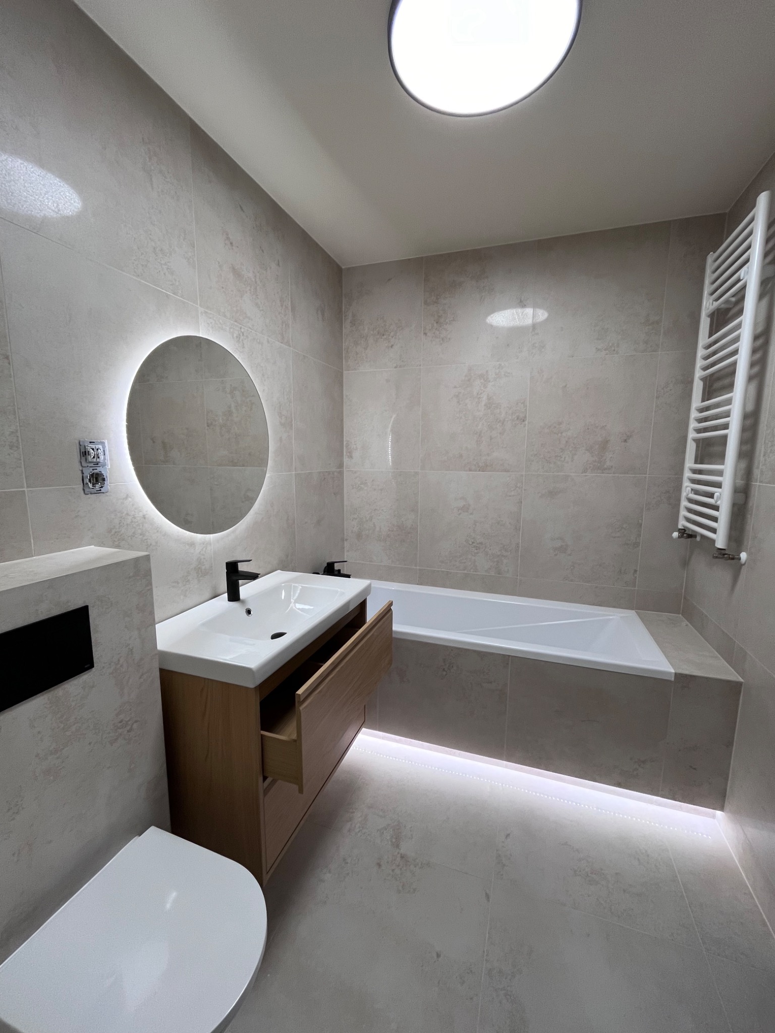 Nowoczesna łazienka z beżowymi płytkami, okrągłym lustrem z podświetleniem LED, drewnianą szafką pod umywalką z otwartą szufladą i podświetleniem pod wanną.