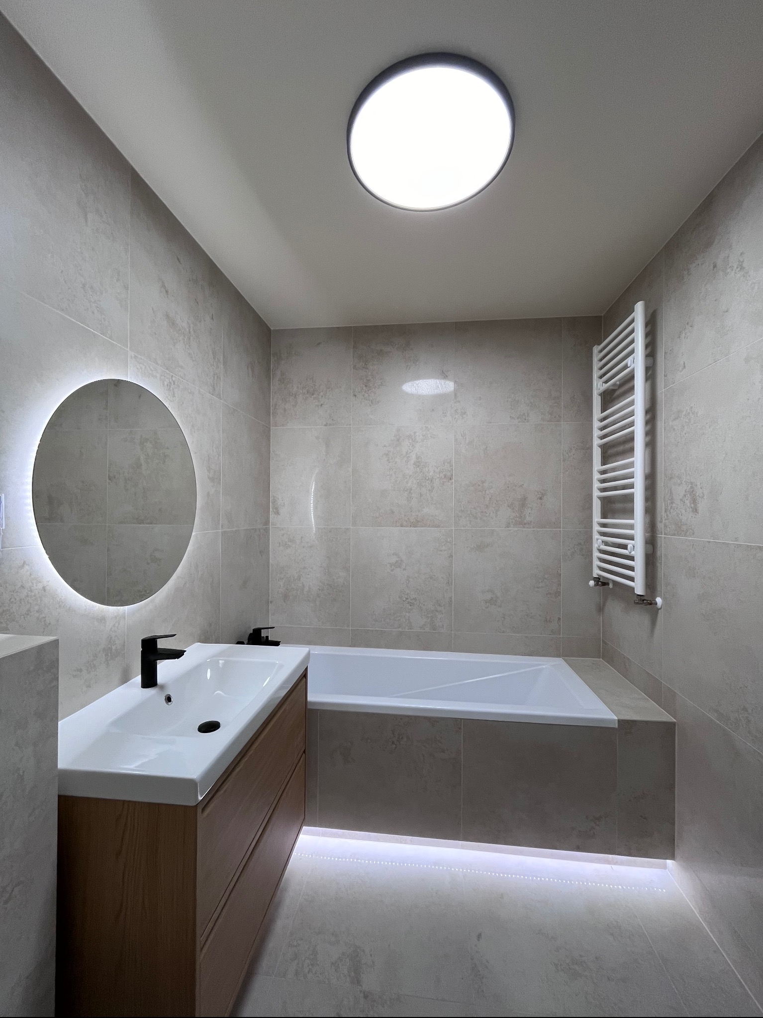 Nowoczesna łazienka z beżowymi płytkami, okrągłym lustrem z podświetleniem LED, drewnianą szafką pod umywalką i podświetlaną wanną.