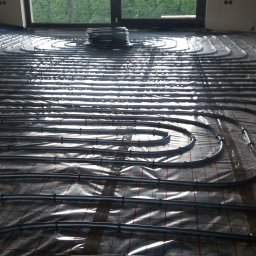 Leonid Aleks - Rozłożone rury systemu ogrzewania podłogowego na folii z siatką w pomieszczeniu z dużym oknem, widoczny zwój rur w tle.