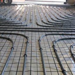 Leonid Aleks - Szare rury systemu ogrzewania podłogowego ułożone równolegle na folii z nadrukowaną siatką, zabezpieczone czarnymi uchwytami, widoczne kartony w tle.