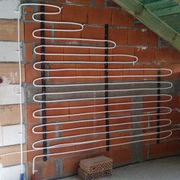 Leonid Aleks - Montaż rur ogrzewania podłogowego na ścianie z cegły dziurawki, widoczne uchwyty mocujące i fragment instalacji elektrycznej przy oknie.