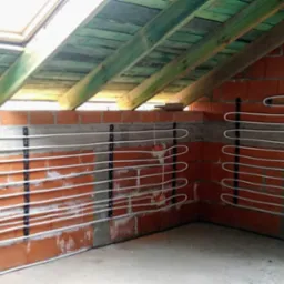Montaż rur w systemie ogrzewania podłogowego na ścianach poddasza ze skosem, widoczne uchwyty mocujące i fragment instalacji hydraulicznej