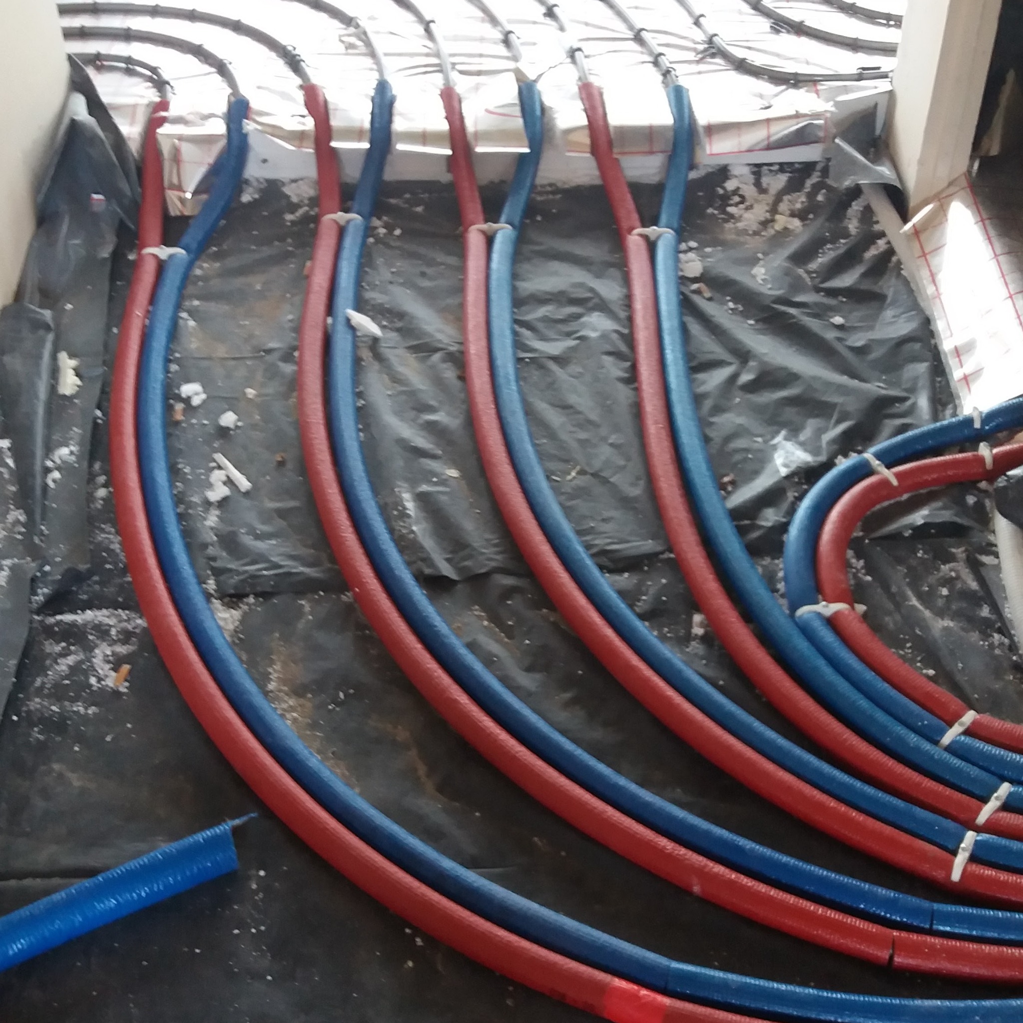 Rozłożone rury ogrzewania podłogowego w kolorach czerwonym i niebieskim, widoczne mocowania do izolacji, czarna folia zabezpieczająca.