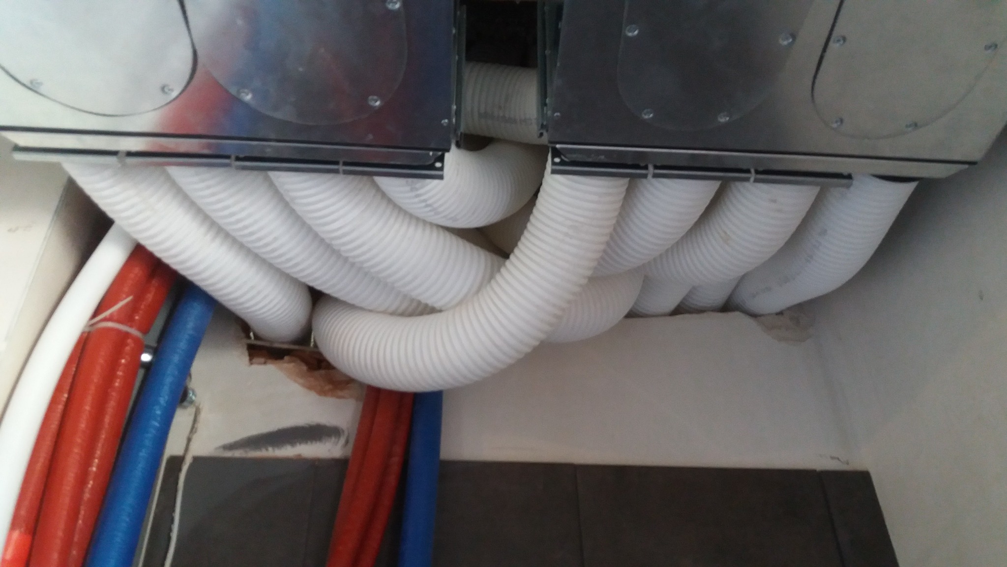 Widok od dołu na podwieszony system wentylacyjny z licznymi białymi, elastycznymi przewodami wentylacyjnymi, oraz fragment instalacji wodnej z rurami w kolorach czerwonym i niebieskim, zamontowany...