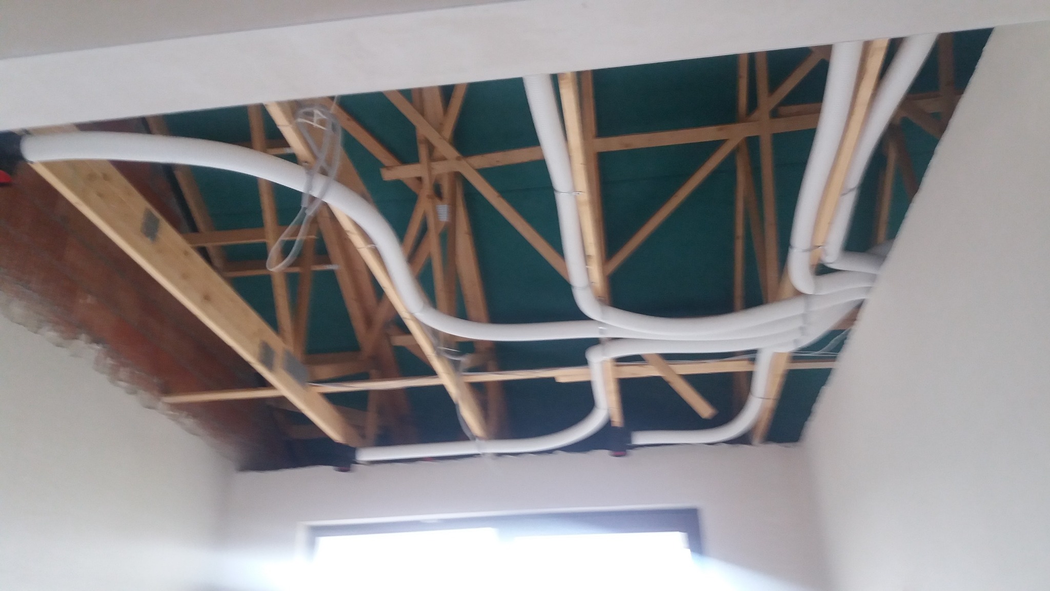 Białe rury wentylacyjne poprowadzone w przestrzeni pod stropem, widoczne drewniane elementy konstrukcji dachu i zielona płyta, częściowo wykończone ściany.