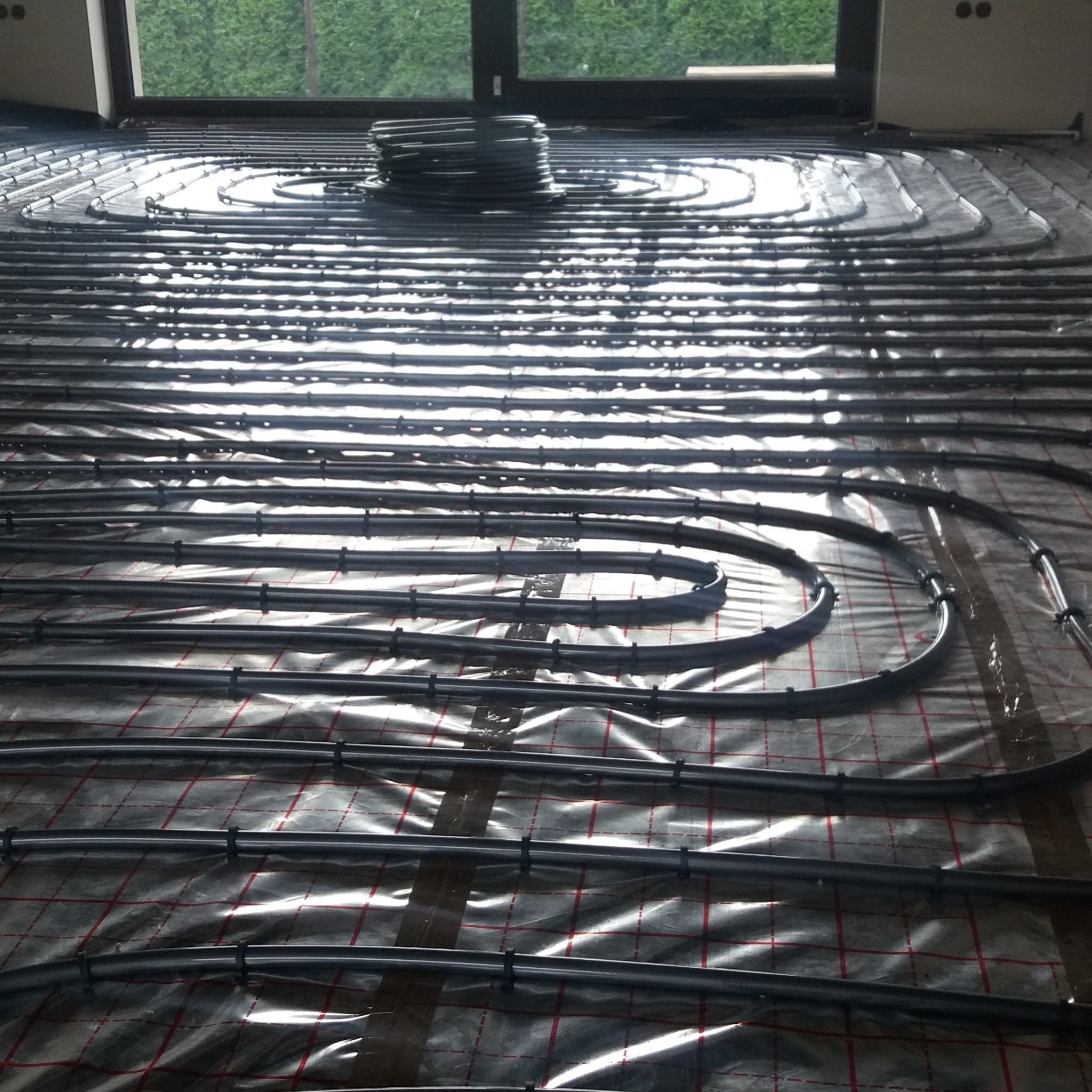 Rozłożone rury systemu ogrzewania podłogowego na folii z siatką w pomieszczeniu z dużym oknem, widoczny zwój rur w tle.