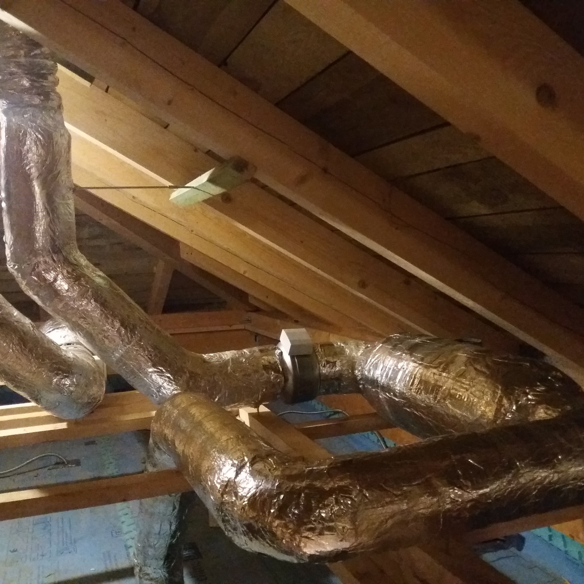 Elastyczne, izolowane przewody wentylacyjne zamontowane pod drewnianą konstrukcją dachu na poddaszu.