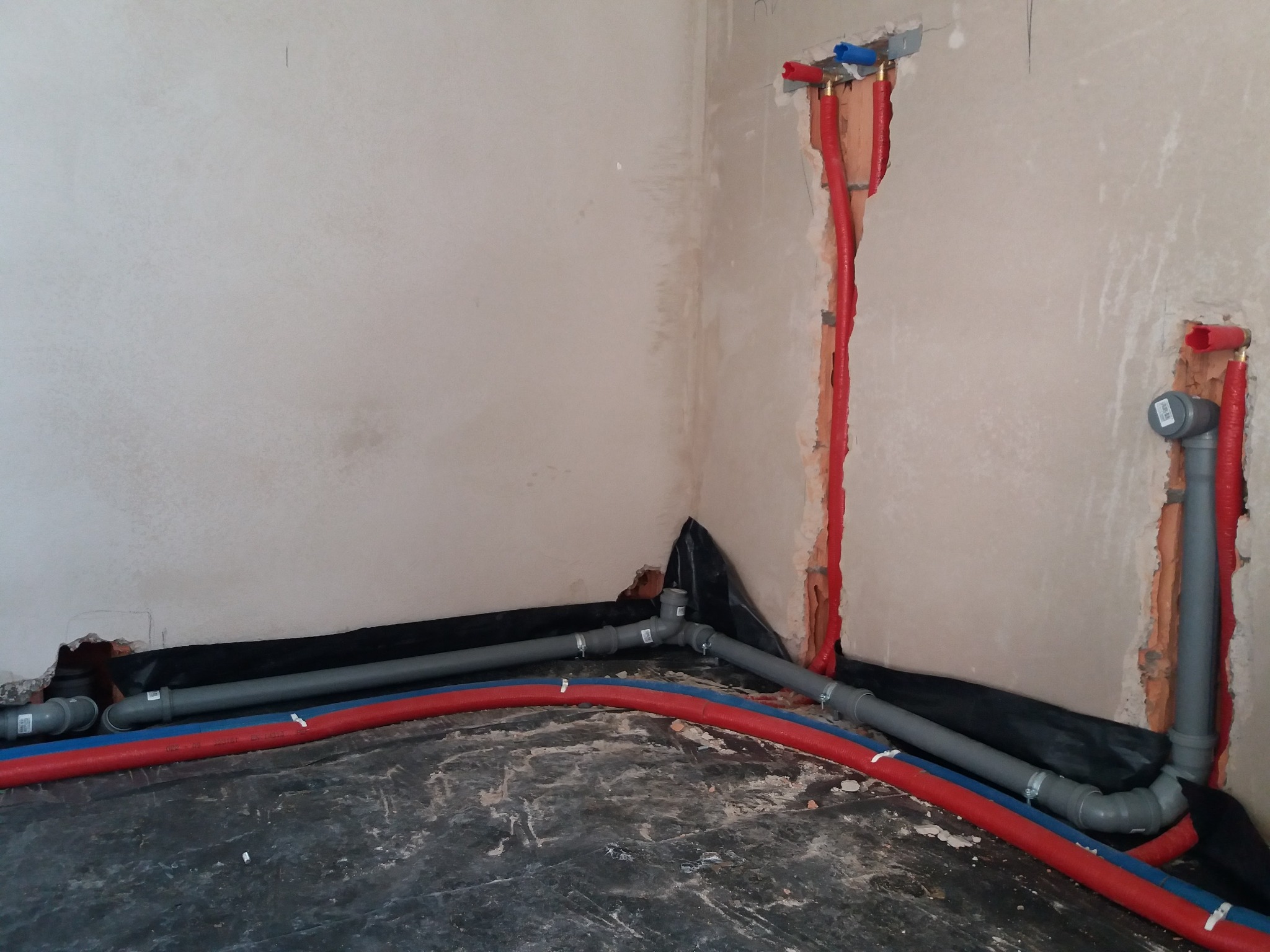 Instalacja wodno-kanalizacyjna w trakcie montażu: rury PEX czerwone i niebieskie oraz szare rury kanalizacyjne ułożone na czarnej folii izolacyjnej, bruzdy w ścianach z wyprowadzonymi rurami.