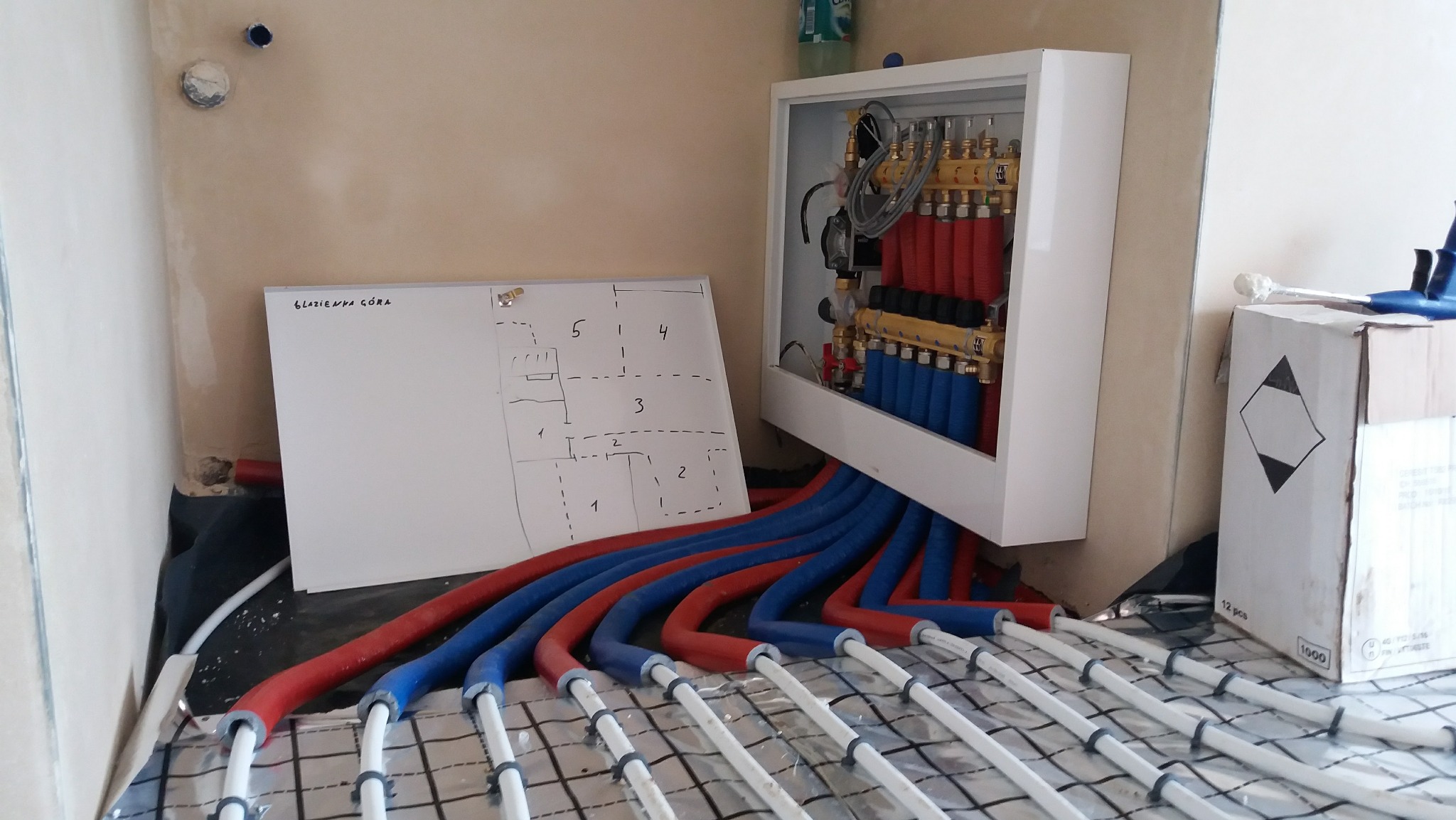 Rozdzielacz ogrzewania podłogowego w białej obudowie z podłączonymi rurami czerwonymi i niebieskimi, widoczny schemat instalacji na tablicy suchościeralnej, całość na folii izolacyjnej...