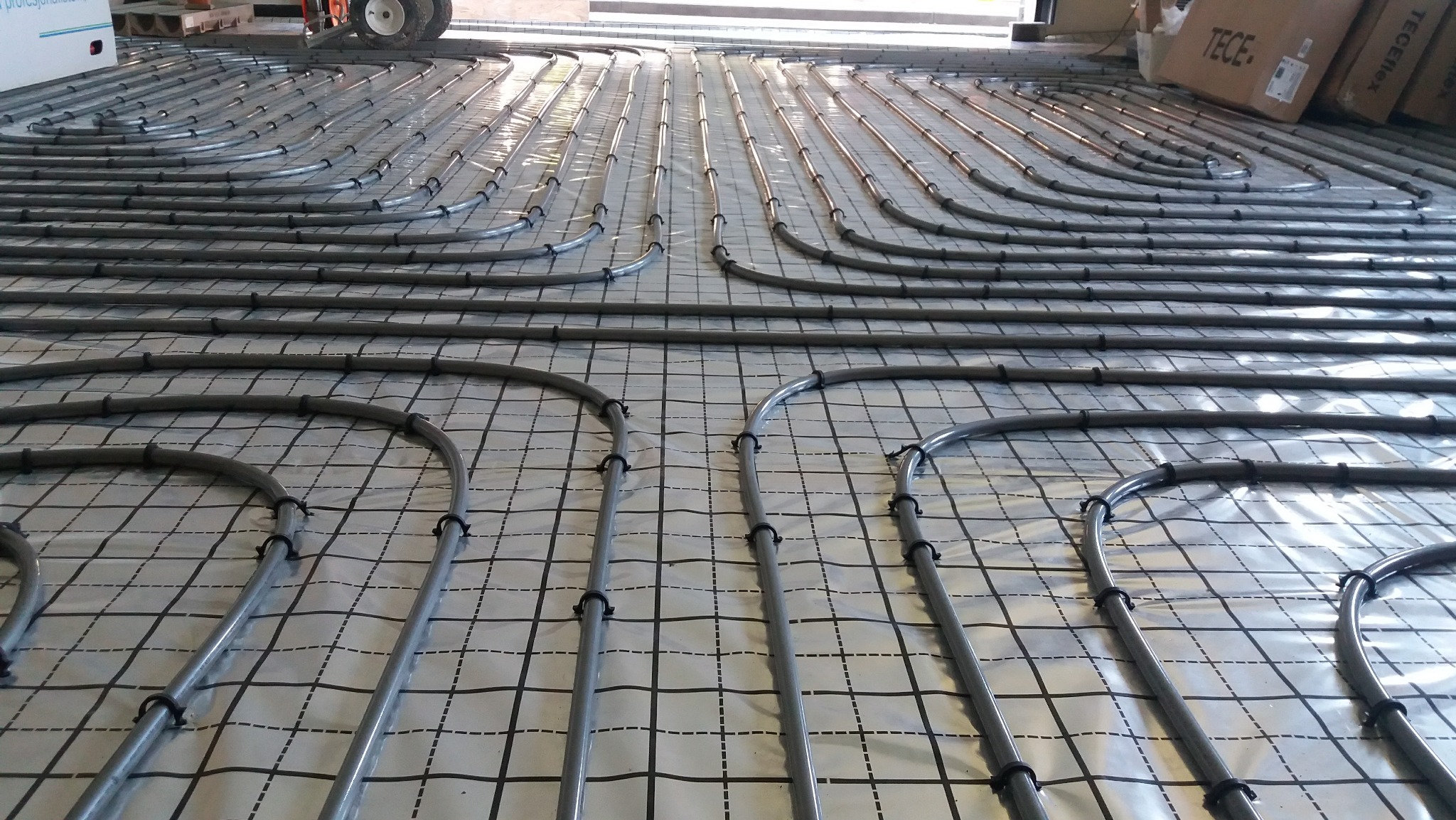 Szare rury systemu ogrzewania podłogowego ułożone równolegle na folii z nadrukowaną siatką, zabezpieczone czarnymi uchwytami, widoczne kartony w tle.