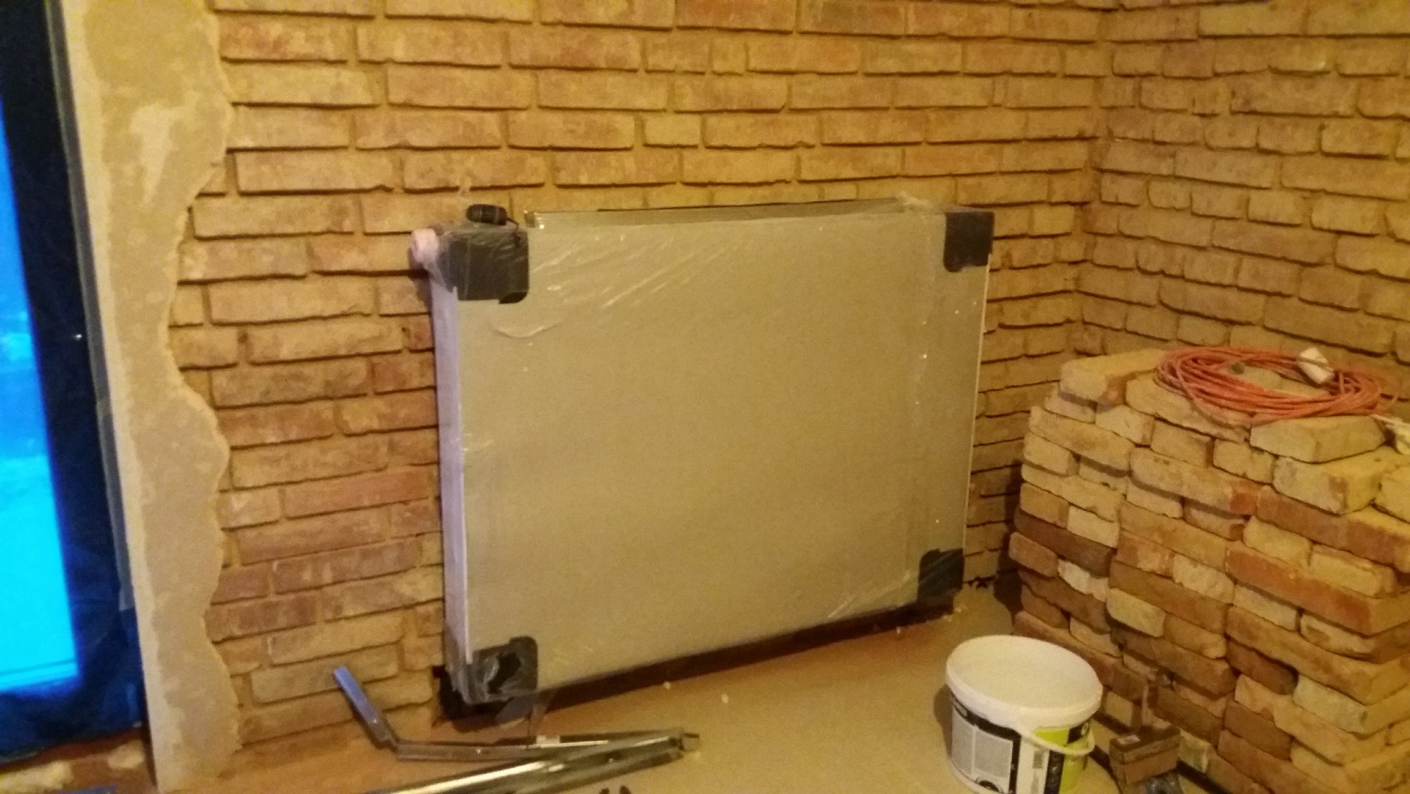 Nowy grzejnik panelowy, owinięty folią ochronną, zamontowany na ścianie z cegły klinkierowej, obok wiadro z farbą i zwój przewodu elektrycznego