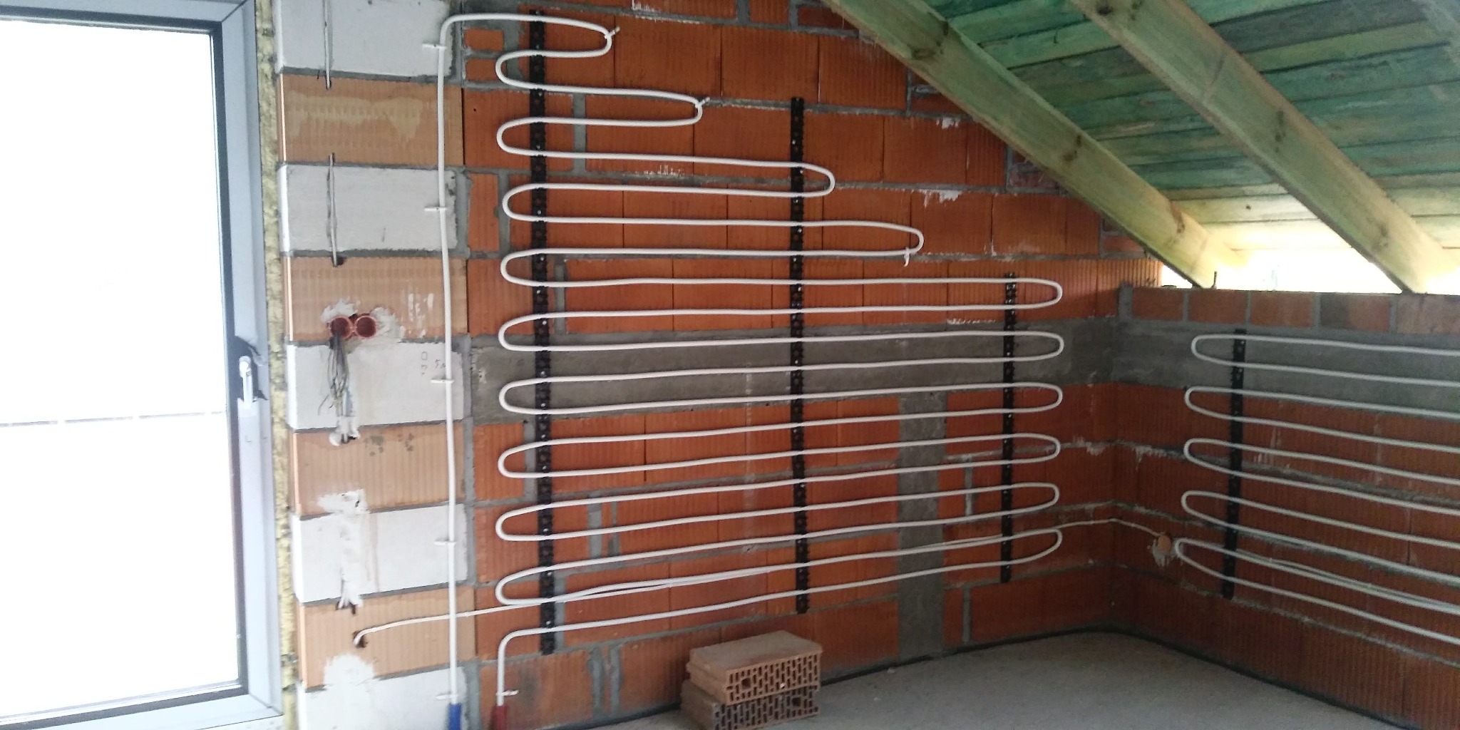 Montaż rur ogrzewania podłogowego na ścianie z cegły dziurawki, widoczne uchwyty mocujące i fragment instalacji elektrycznej przy oknie.