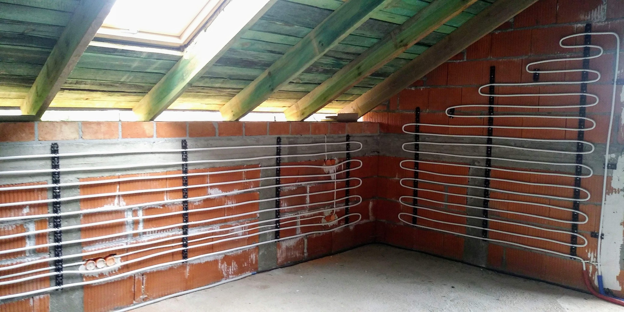 Montaż rur w systemie ogrzewania podłogowego na ścianach poddasza ze skosem, widoczne uchwyty mocujące i fragment instalacji hydraulicznej