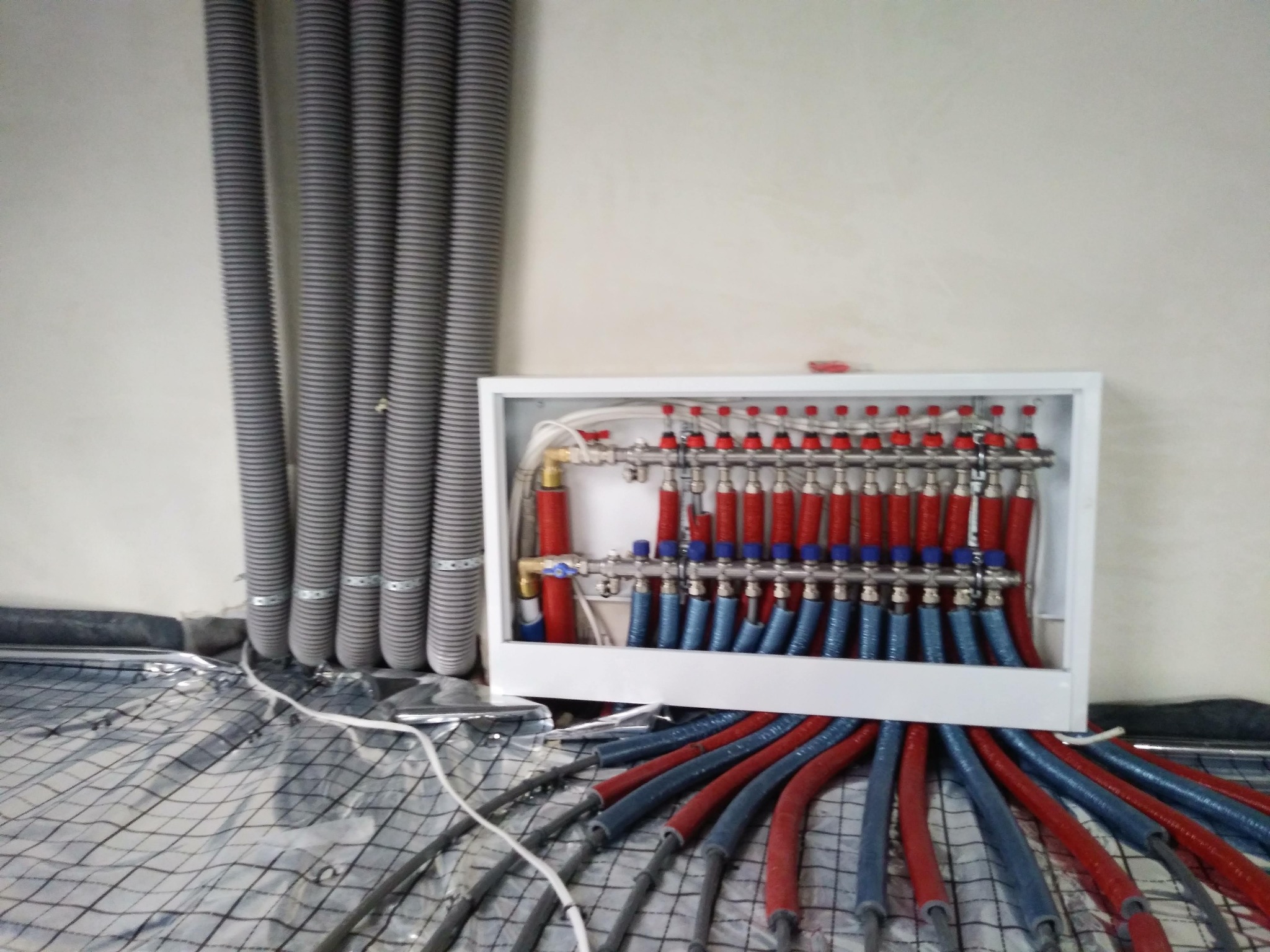 Rozdzielacz ogrzewania podłogowego w białej obudowie, z widocznymi rurami w kolorach czerwonym i niebieskim, ułożony na folii izolacyjnej z kratką, z szarymi rurami wentylacyjnymi po lewej stronie.