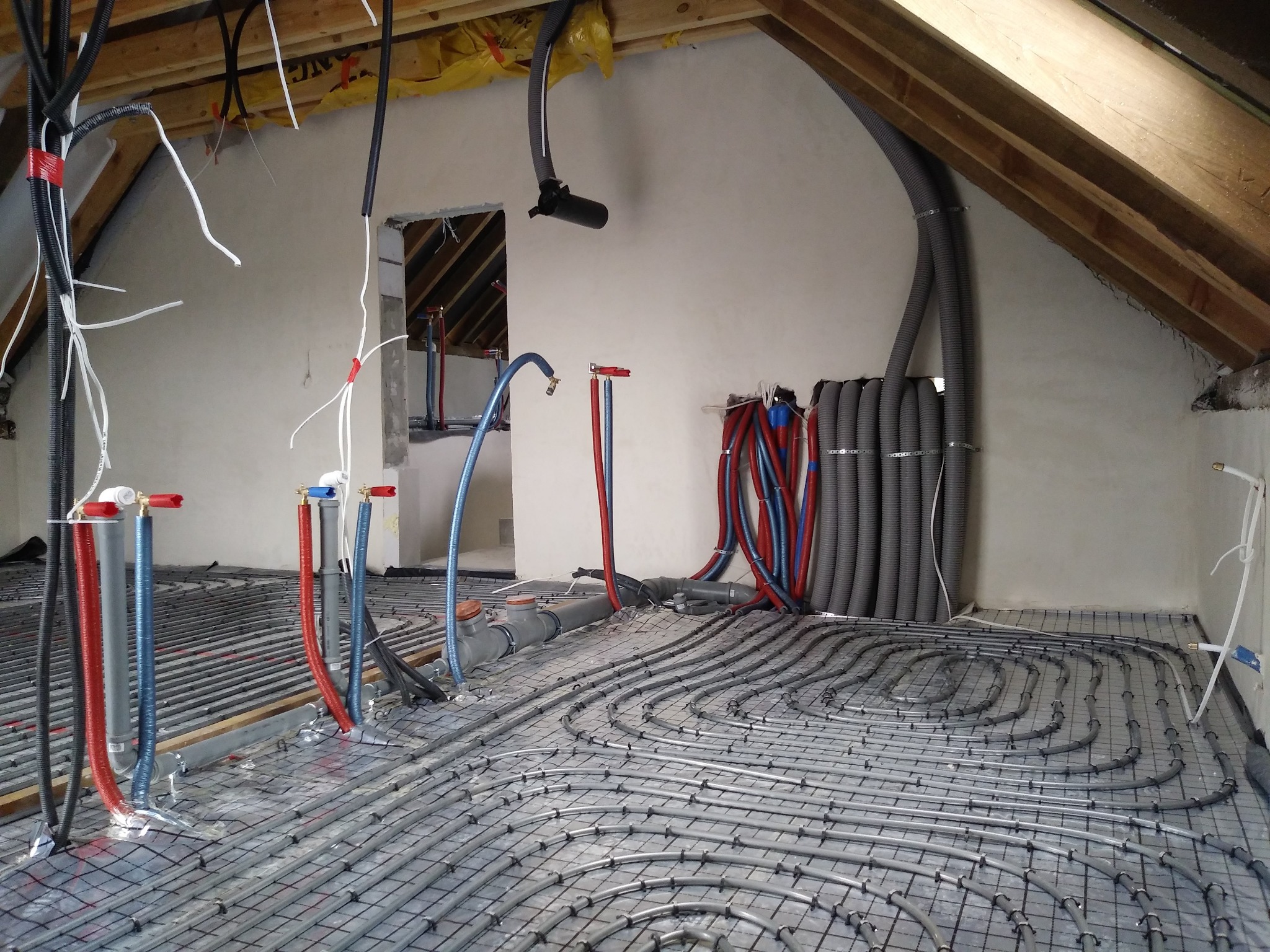 Instalacja ogrzewania podłogowego w trakcie montażu na poddaszu z widocznymi rurami w szarej siatce montażowej oraz elementami instalacji wentylacyjnej pod sufitem.