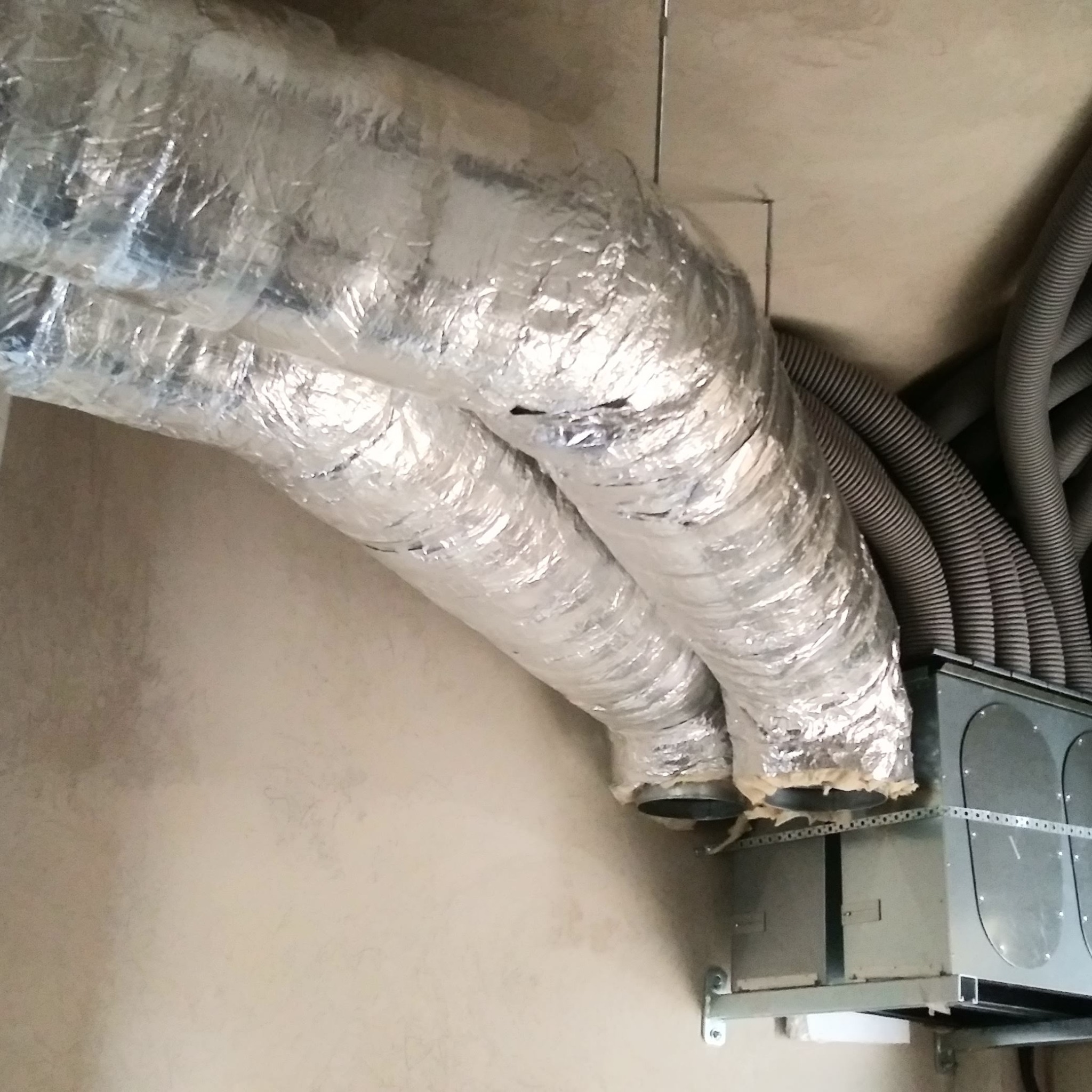 Dwa izolowane przewody wentylacyjne z folii aluminiowej łączą się z centralą wentylacyjną, szare rury karbowane w tle.