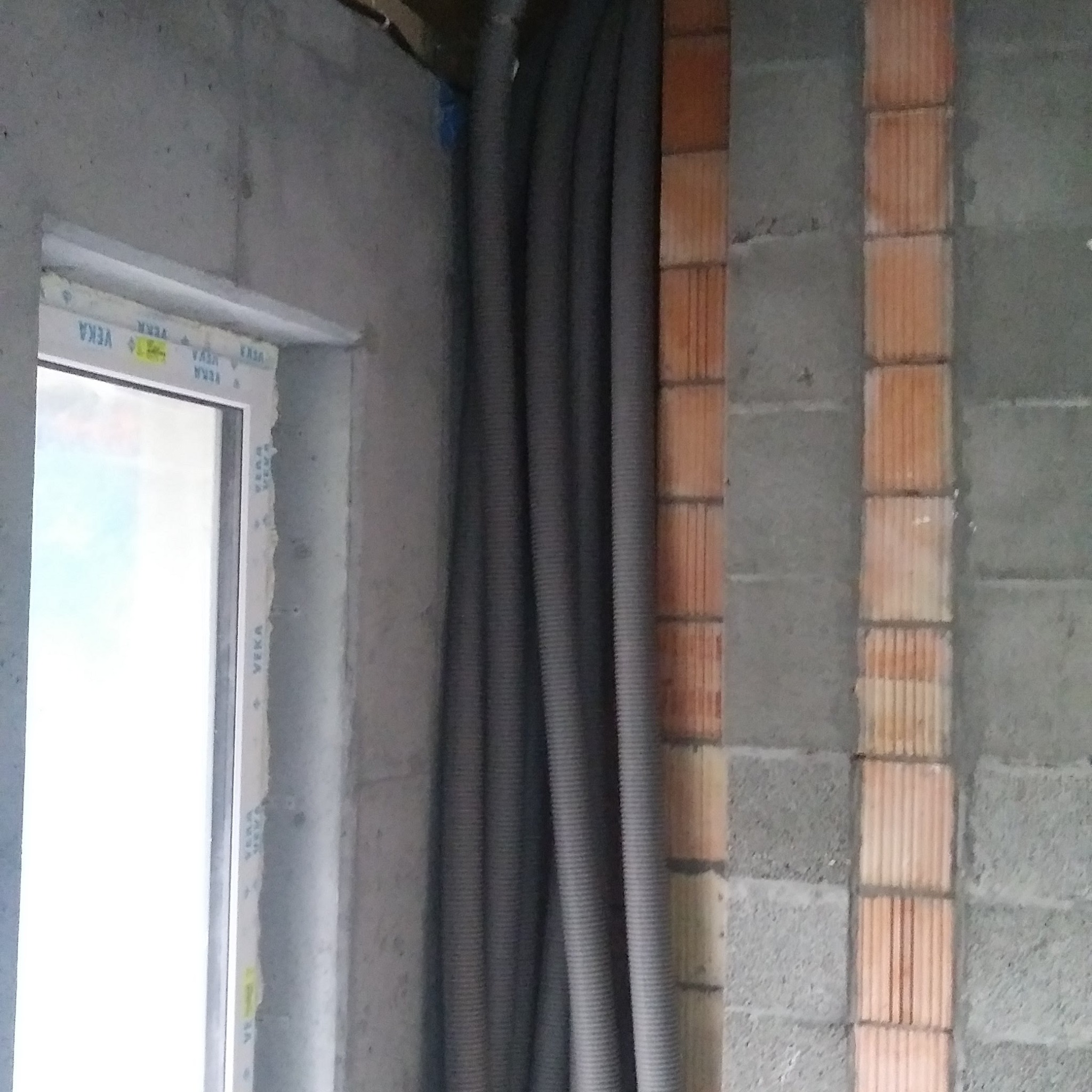 Szare, karbowane rury wentylacyjne zwisają w surowym wnętrzu, widoczne obok okna z białymi ramami i fragmentu ściany z czerwonej cegły.