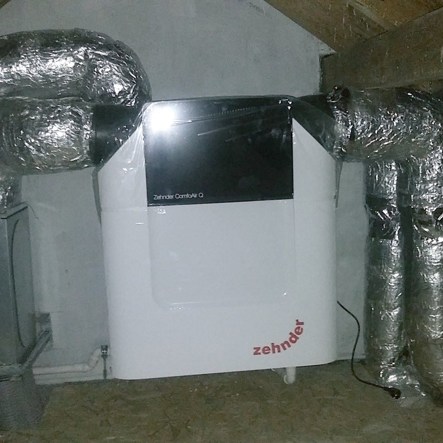 Biała jednostka centralna Zehnder ComfoAir Q z podłączonymi, zaizolowanymi przewodami wentylacyjnymi, umieszczona w pomieszczeniu z betonową ścianą i drewnianym stropem.