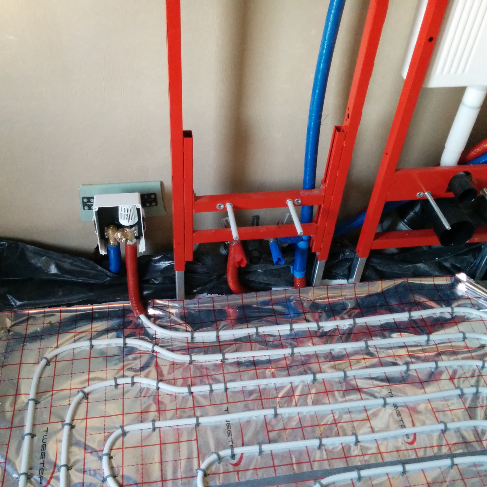 Montaż ogrzewania podłogowego: rury grzewcze ułożone na folii izolacyjnej z siatką, widoczne elementy instalacji sanitarnej w tle, czerwona rama mocująca i regulator temperatury na ścianie.