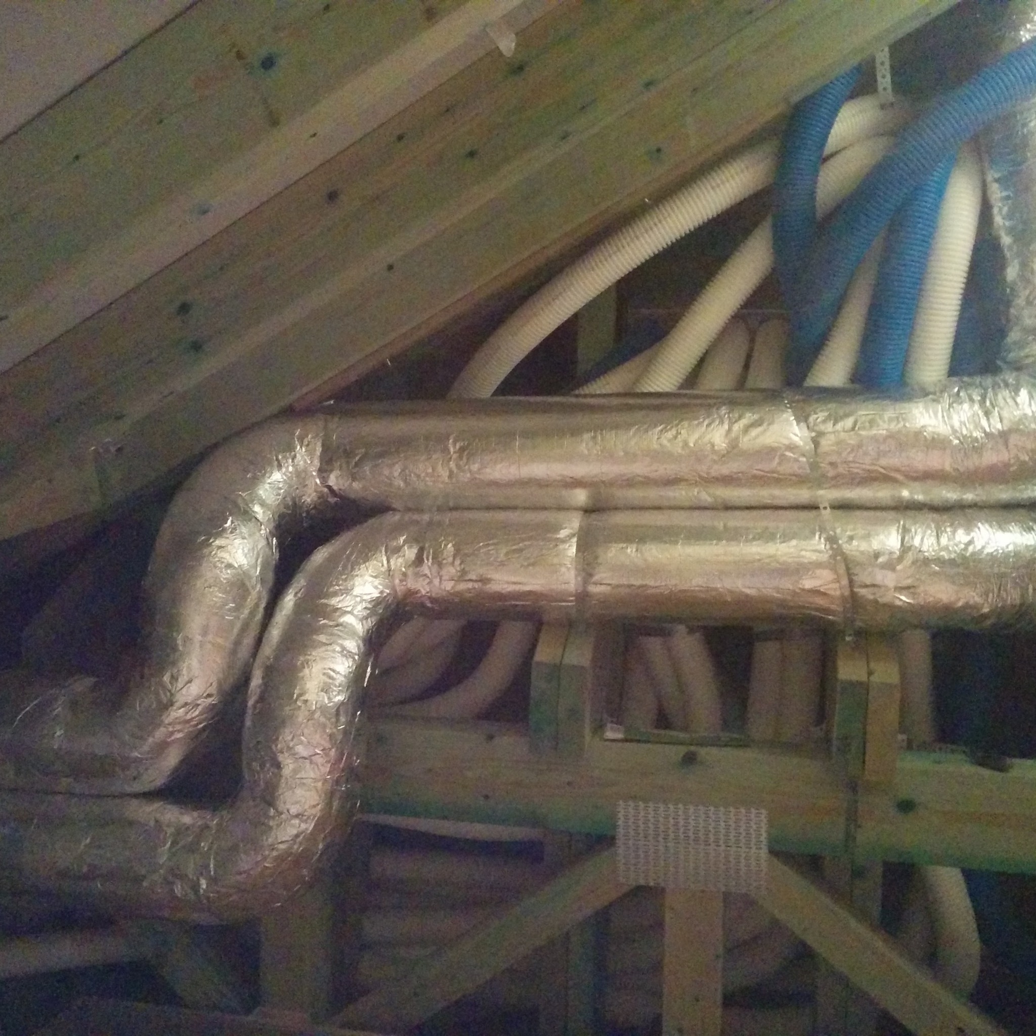 Fragment poddasza z widocznymi izolowanymi przewodami wentylacyjnymi, niektóre z nich w kolorze niebieskim, zainstalowanymi między drewnianą konstrukcją dachu.