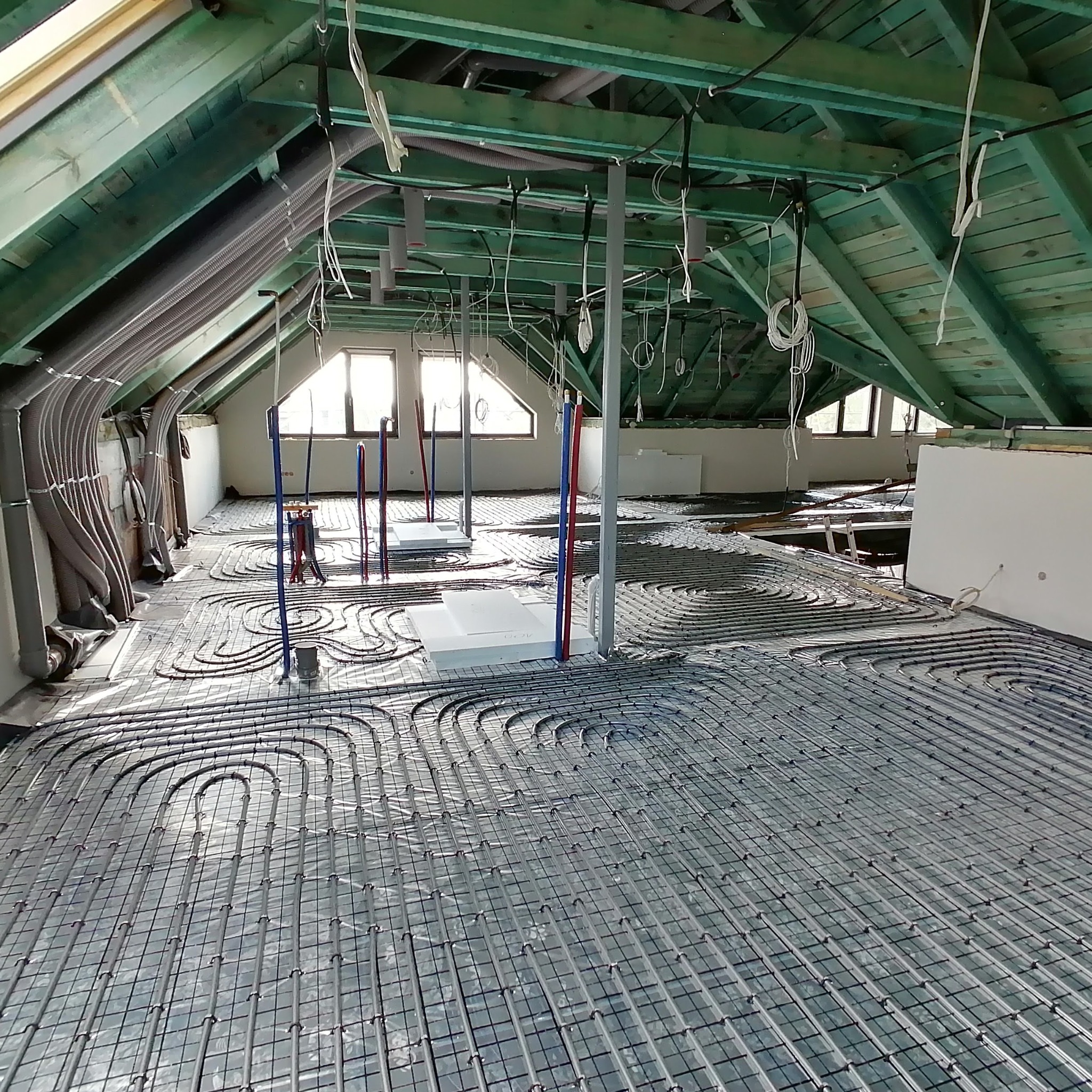 Instalacja ogrzewania podłogowego w trakcie montażu na poddaszu, widoczne rury grzewcze ułożone w pętle na folii izolacyjnej i siatce, wystające pionowe rury doprowadzające, surowy stan wykończenia...