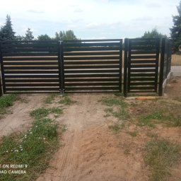 RENEX Ireneusz Kwiatkowski - Nowoczesne, czarne ogrodzenie panelowe z poziomymi prętami, zamontowane na posesji z widoczną ziemią i trawą, w tle drzewa i dom.