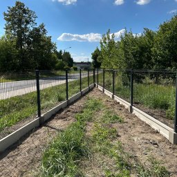 RENEX Ireneusz Kwiatkowski - Montaż ogrodzenia panelowego z podmurówką betonową na działce z widoczną drogą w tle, w słoneczny dzień.