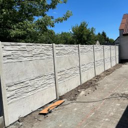 RENEX Ireneusz Kwiatkowski - Długi, betonowy płot z panelami o fakturze kamienia, oddzielający posesję od chodnika z kostki brukowej, widok za dnia, z zielenią drzew w tle.