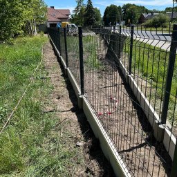 RENEX Ireneusz Kwiatkowski - Nowo postawione, czarne ogrodzenie panelowe na betonowej podmurówce, widoczne z boku wzdłuż działki, z trawą po obu stronach i rozciągniętą czerwoną taśmą ostrzegawczą.