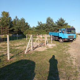 RENEX Ireneusz Kwiatkowski - W trakcie montażu ogrodzenia z betonowych słupków i siatki, widoczny niebieski samochód ciężarowy MAN oraz cień osoby robiącej zdjęcie.