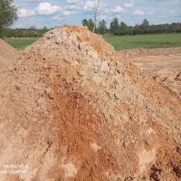 RENEX Ireneusz Kwiatkowski - Kupa piasku i ziemi na placu budowy, z widocznym wykopem w tle i osobą siedzącą na szczycie innej pryzmy ziemi.