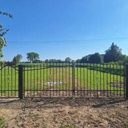 RENEX Ireneusz Kwiatkowski - Czarna, metalowa brama dwuskrzydłowa z ozdobnymi elementami, zamontowana na posesji z widokiem na trawnik i zieleń, z kwitnącym krzewem po lewej stronie i słupami energetycznymi w tle.