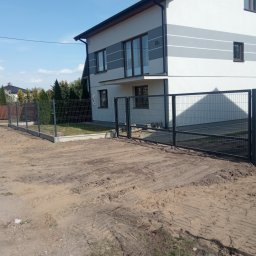 RENEX Ireneusz Kwiatkowski - Nowoczesny dom z szarą elewacją i nowo zamontowanym, czarnym ogrodzeniem panelowym z podmurówką, widok na nierówny teren przed posesją.