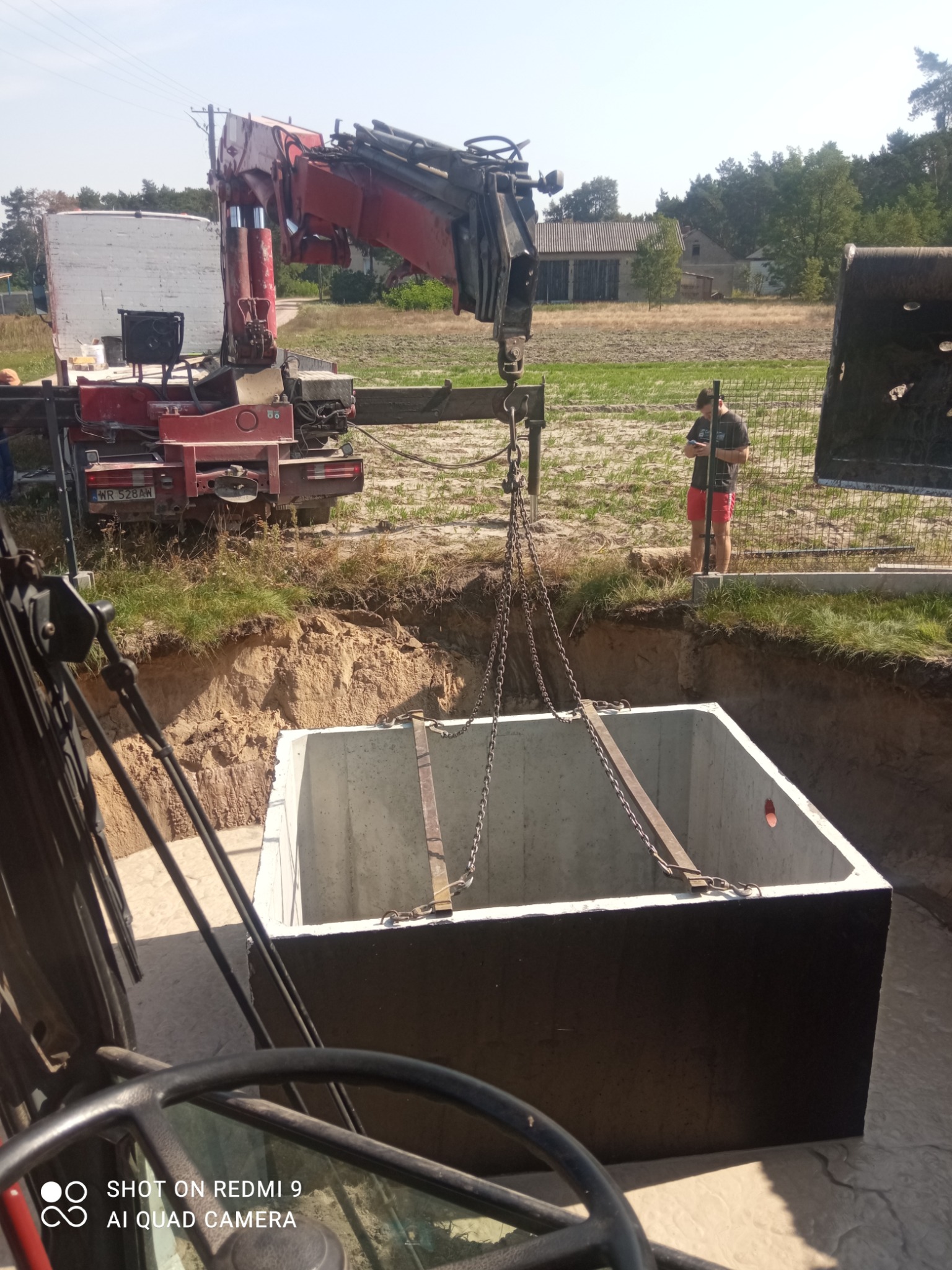 Ciężki, betonowy zbiornik opuszczany do wykopu za pomocą dźwigu na tle krajobrazu wiejskiego.