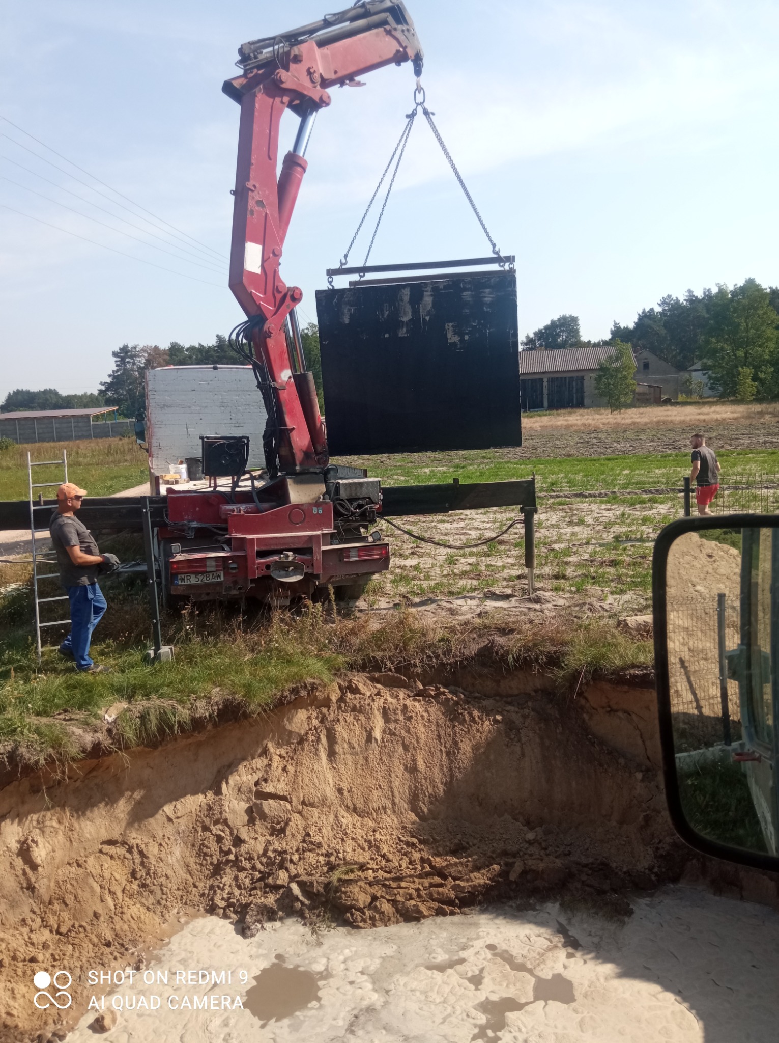Czerwony dźwig samochodowy opuszcza czarny, prostokątny element do wykopu wypełnionego wodą na działce z budynkami w tle, nadzór pracownika w pomarańczowej czapce.