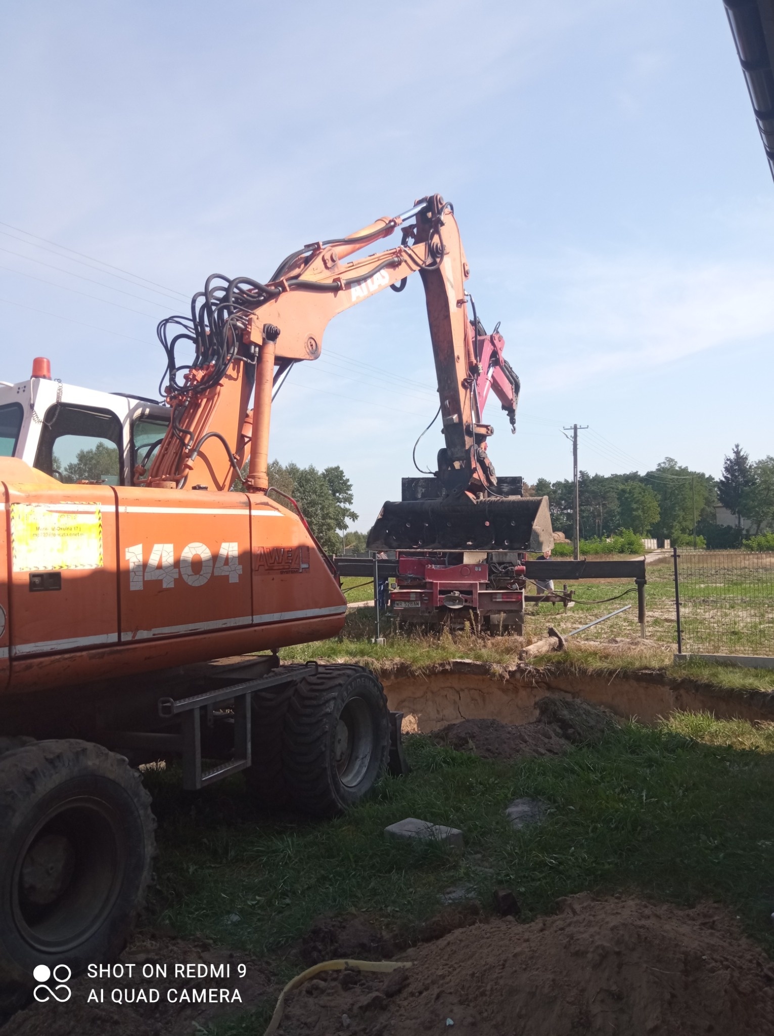 Pomarańczowa koparka Atlas 1404 na placu budowy, ramię maszyny nad wykopem, w tle widoczny samochód ciężarowy z zamontowanym urządzeniem do wbijania pali.