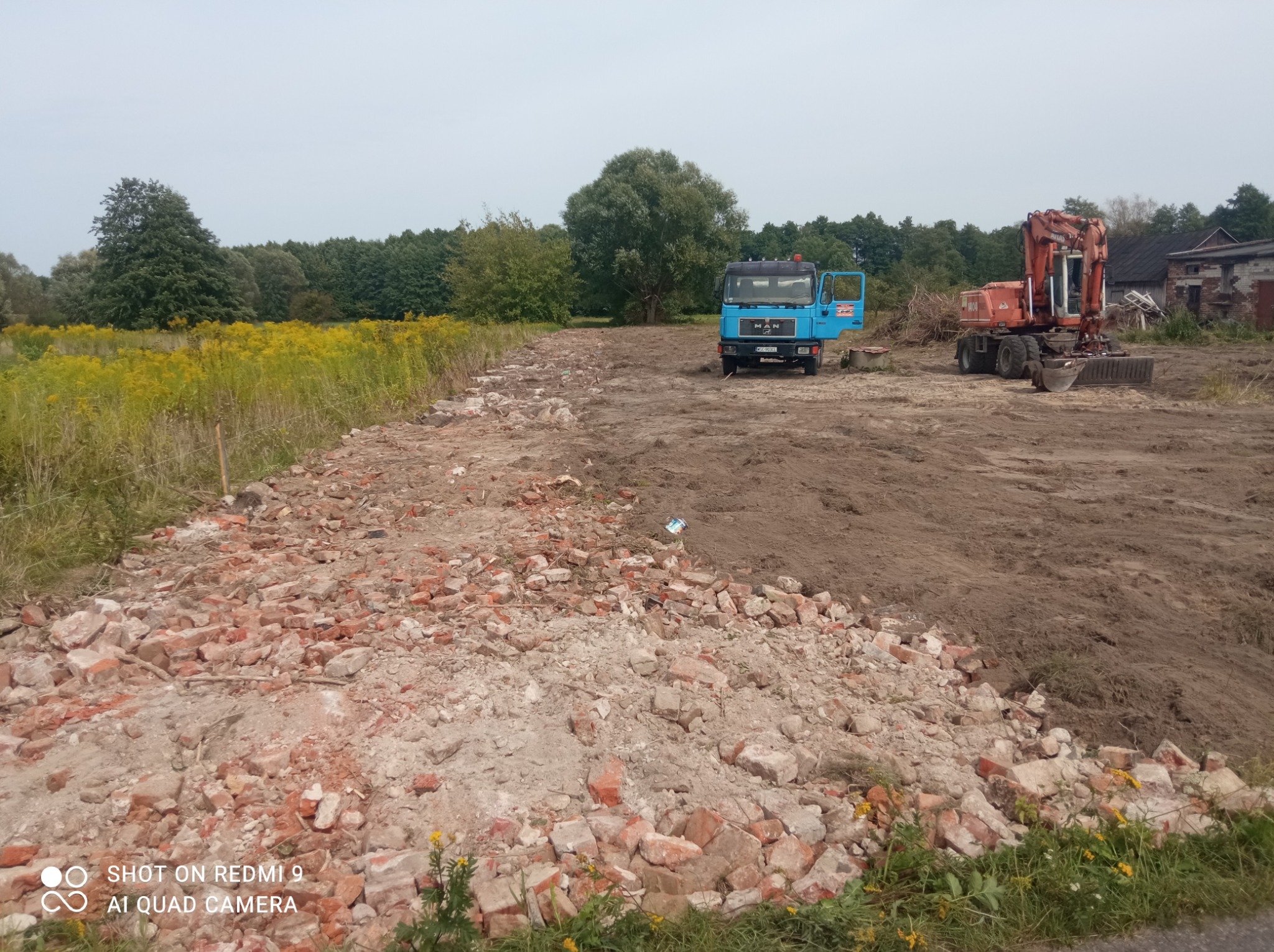 Nierówny teren z gruzem ceglanym, przygotowywany pod wyrównanie, z widocznym niebieskim samochodem ciężarowym MAN i pomarańczową koparką w tle, oraz polami żółtych kwiatów i drzewami.