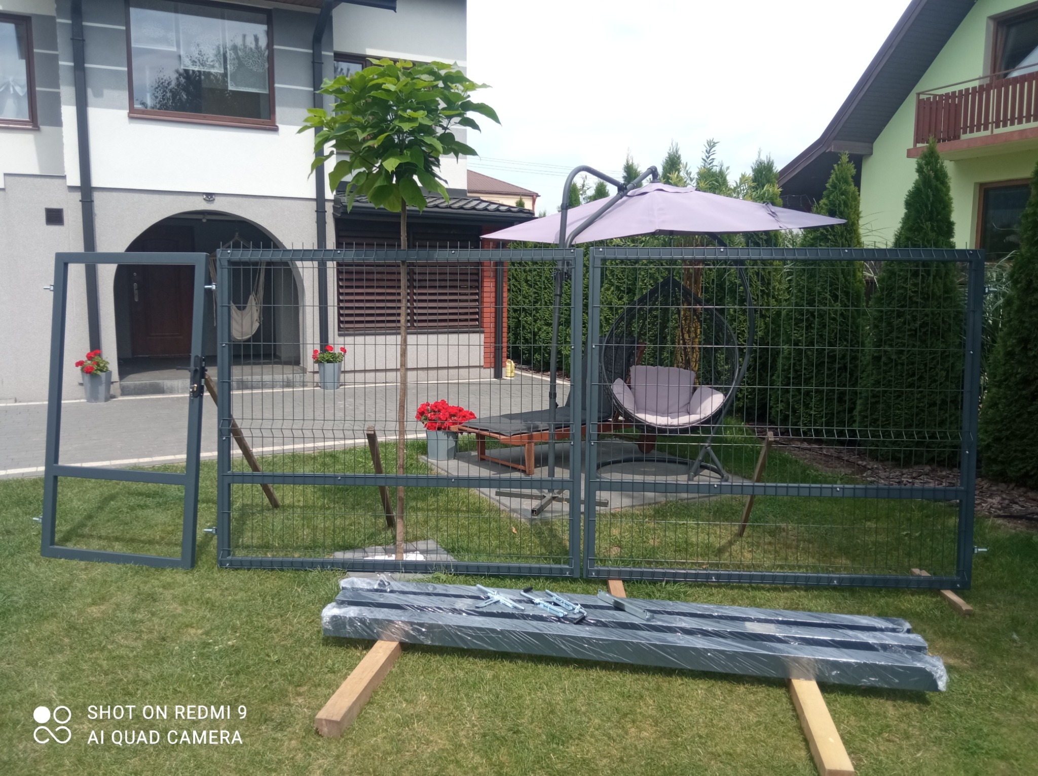 Częściowo zamontowane szare ogrodzenie panelowe na trawniku przed domem, z elementami do montażu w folii na drewnianych legarach, widoczny fragment furtki i sąsiednie budynki w tle.