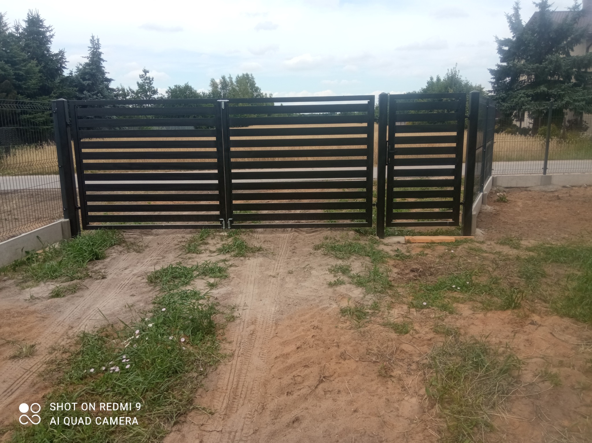 Nowoczesne, czarne ogrodzenie panelowe z poziomymi prętami, zamontowane na posesji z widoczną ziemią i trawą, w tle drzewa i dom.