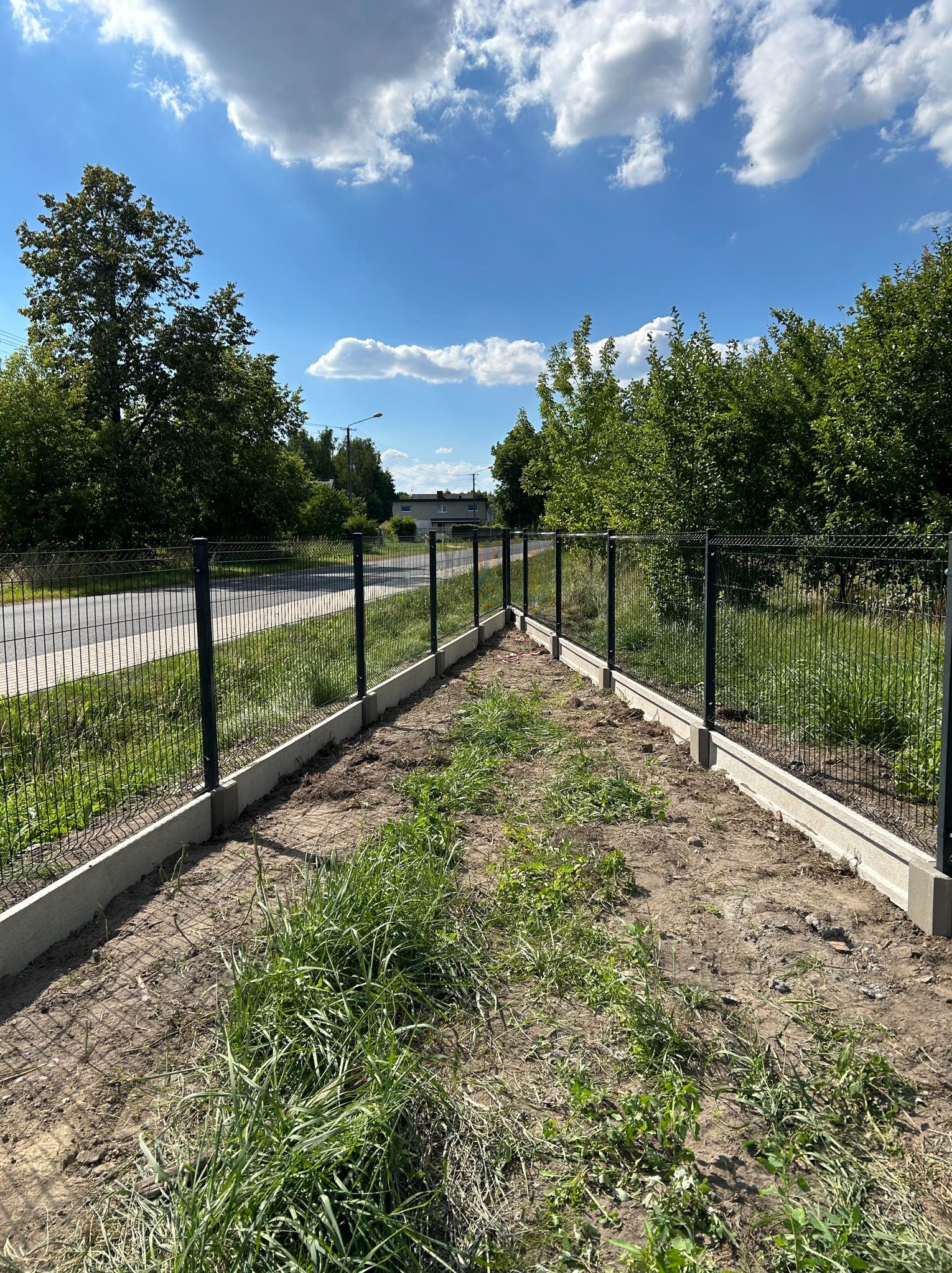 Montaż ogrodzenia panelowego z podmurówką betonową na działce z widoczną drogą w tle, w słoneczny dzień.