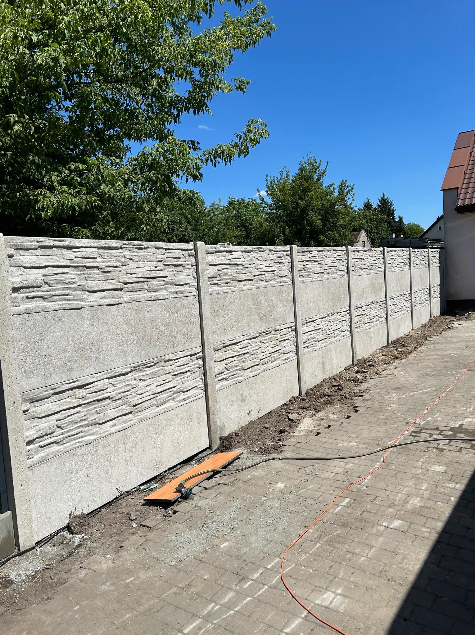 Długi, betonowy płot z panelami o fakturze kamienia, oddzielający posesję od chodnika z kostki brukowej, widok za dnia, z zielenią drzew w tle.